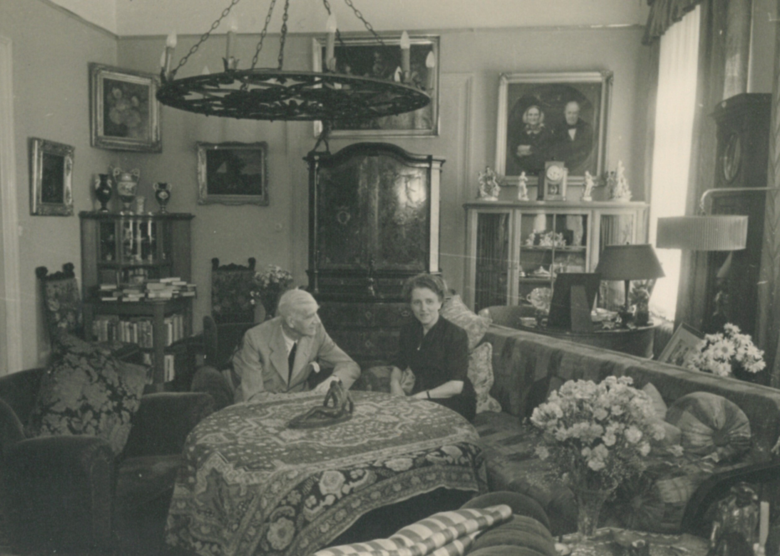 Wohnzimmer mit Eheaar Fievet. Ein älteres Ehepaar sitzt gemeinsam an einem runden Tisch mit einer gemusterten Tischdecke in der Mitte des Raumes. Die Frau sitzt auf einem langen Sofa. Der Mann sitzt auf einem Sessel und schaut die Frau an. Über dem Paar hängt ein runder Kerzenleuchter. Zwei große Blumensträuße stehen im Raum verteilt. Drei Schränke und Regale befinden sich an der Wand im Hintergrund. Fünf verschieden große Gemälde mit Pflanzen oder Menschen hängen an der Wand.