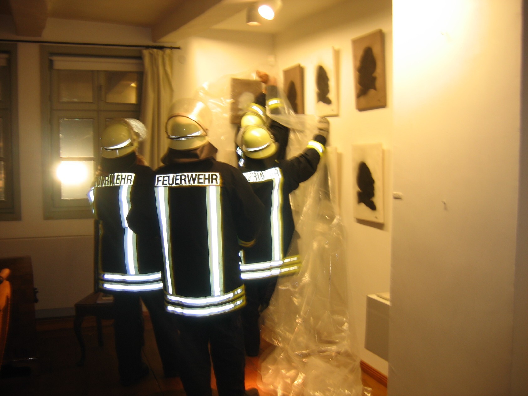 Vier Personen stehen mit dem Rücken zum Betrachtenden. Sie tragen gelbe Helme und schwarze Jacken mit Reflektorstreifen und der Aufschrift Feuerwehr. Die Personen decken an der Wand hängende Scherenschnitte mit einer Folie ab.