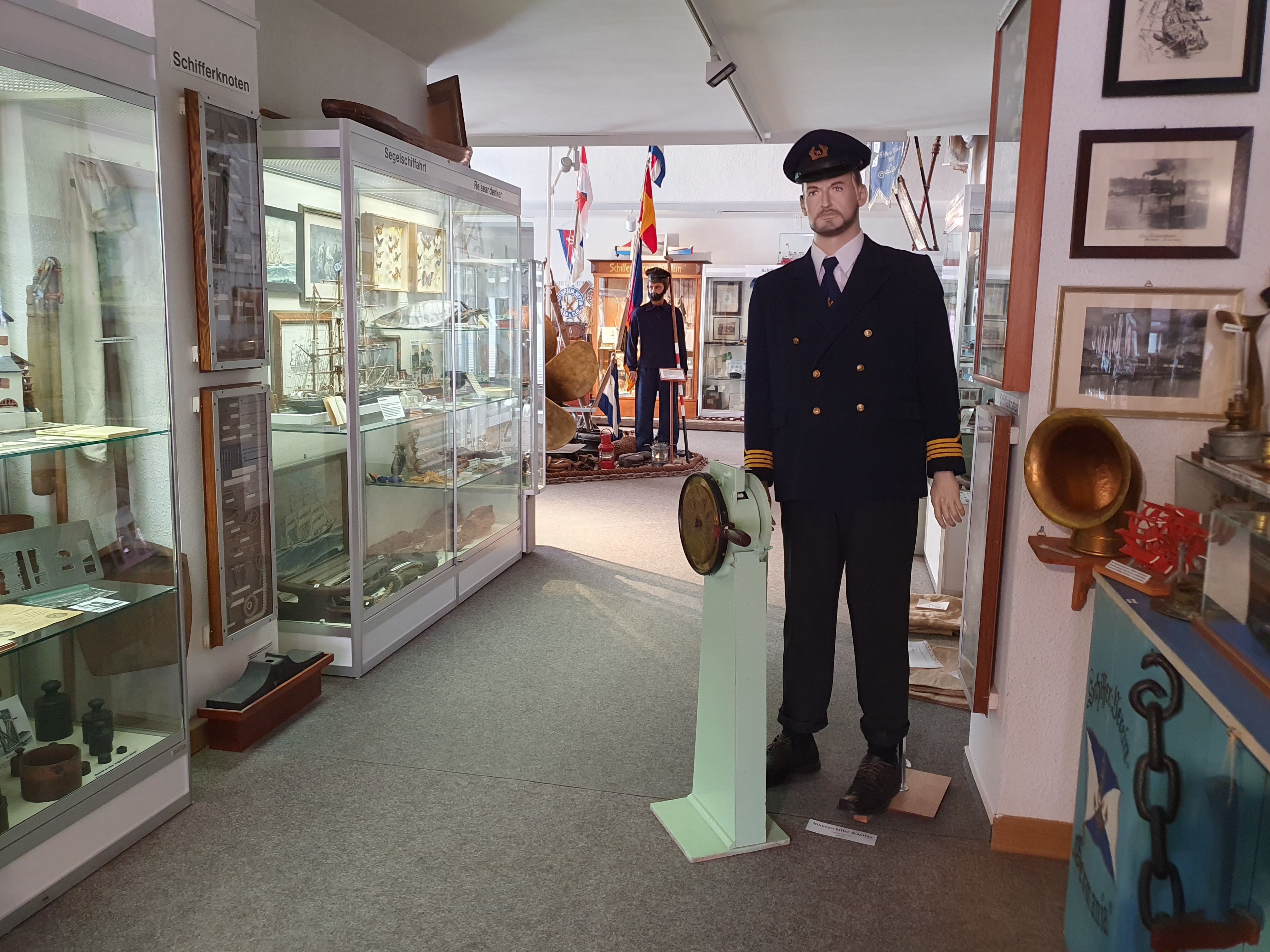 Eine als Kapitän gekleidete menschengroße Puppe steht am Eingang der Ausstellung. Im Hintergrund stehen Vitrinen mit Schiffsmodellen und Gemälden. Ein Matrose umgeben von Flaggen steht dahinter.