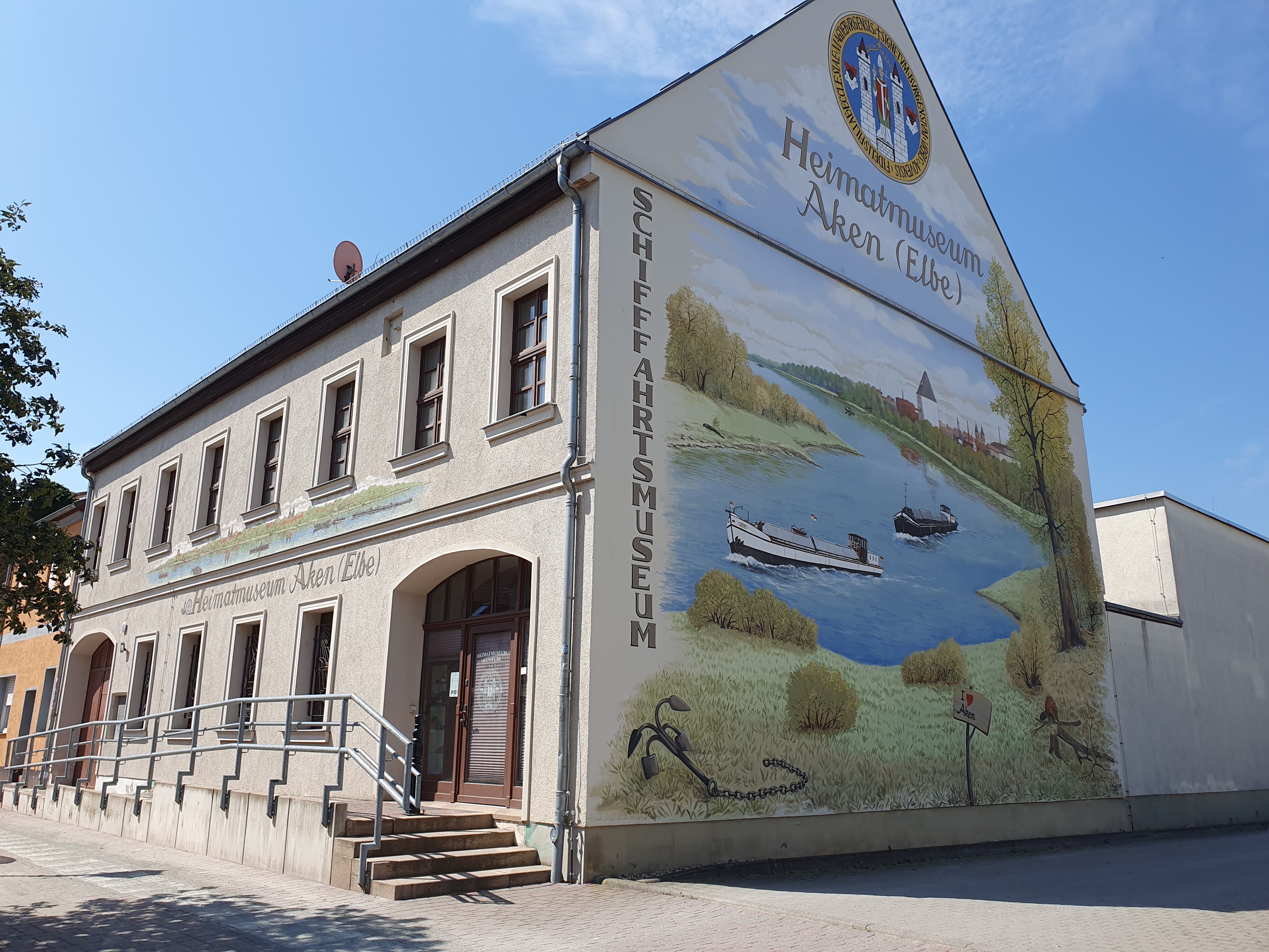 Die Außenfassade des Heimat- und Schifffahrtsmuseums Aken. Auf der bemalten Fassade sind zwei Schiffe auf einem Fluss vom Ufer aus erkennbar.
