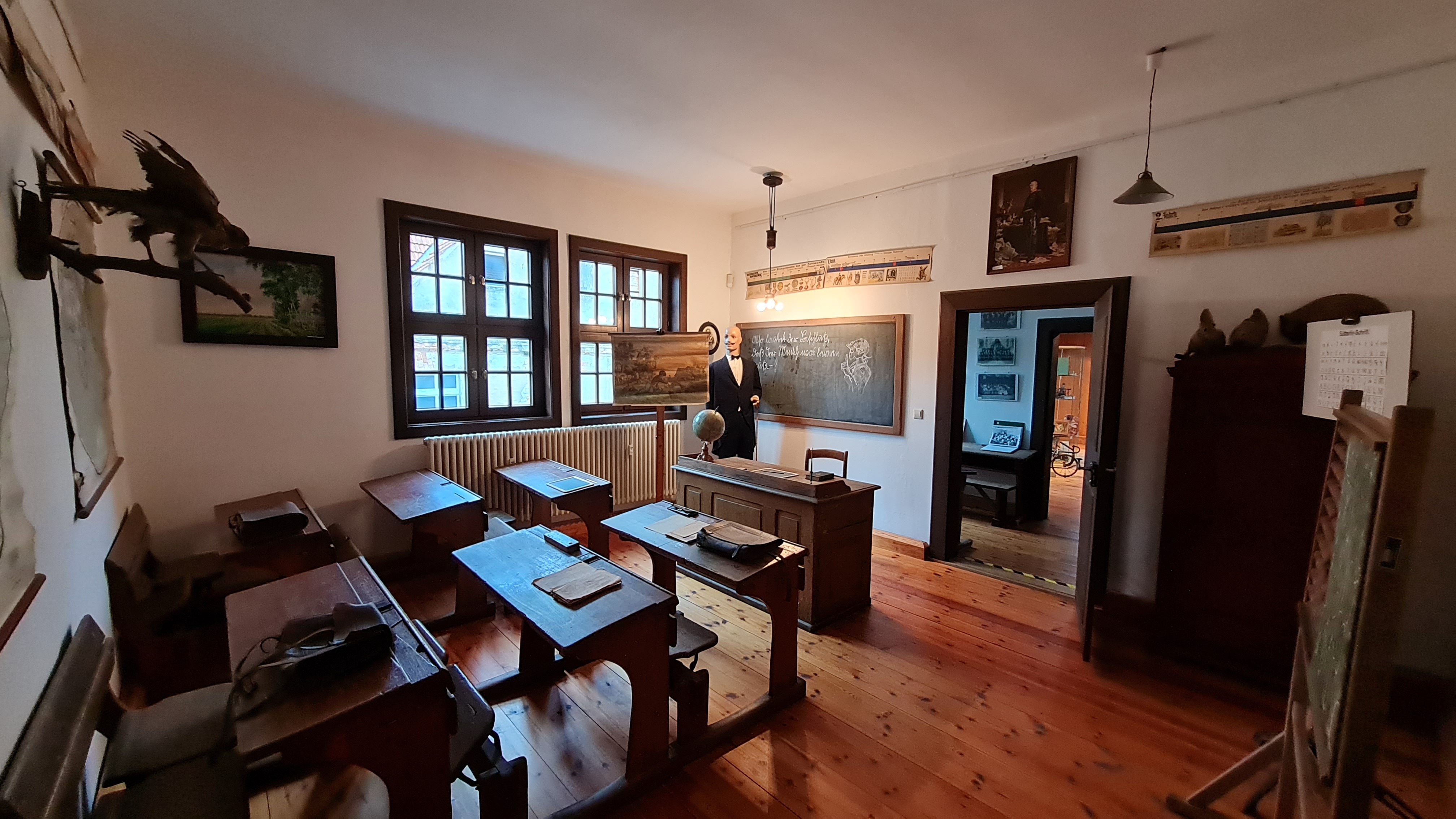 Ein Klassenzimmer wie um 1900 eingerichtet. Sechs Schreibbänke aus dunklem Holz sind auf die Tafel und den Lehrertisch ausgerichtet.