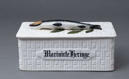 Eine Dose aus weißem Steingut aus dem Jahr 1890. Auf der Dose steht "Marinierte Heringe" geschrieben und ein Hering aus Porzellan liegt auf dem Deckel.