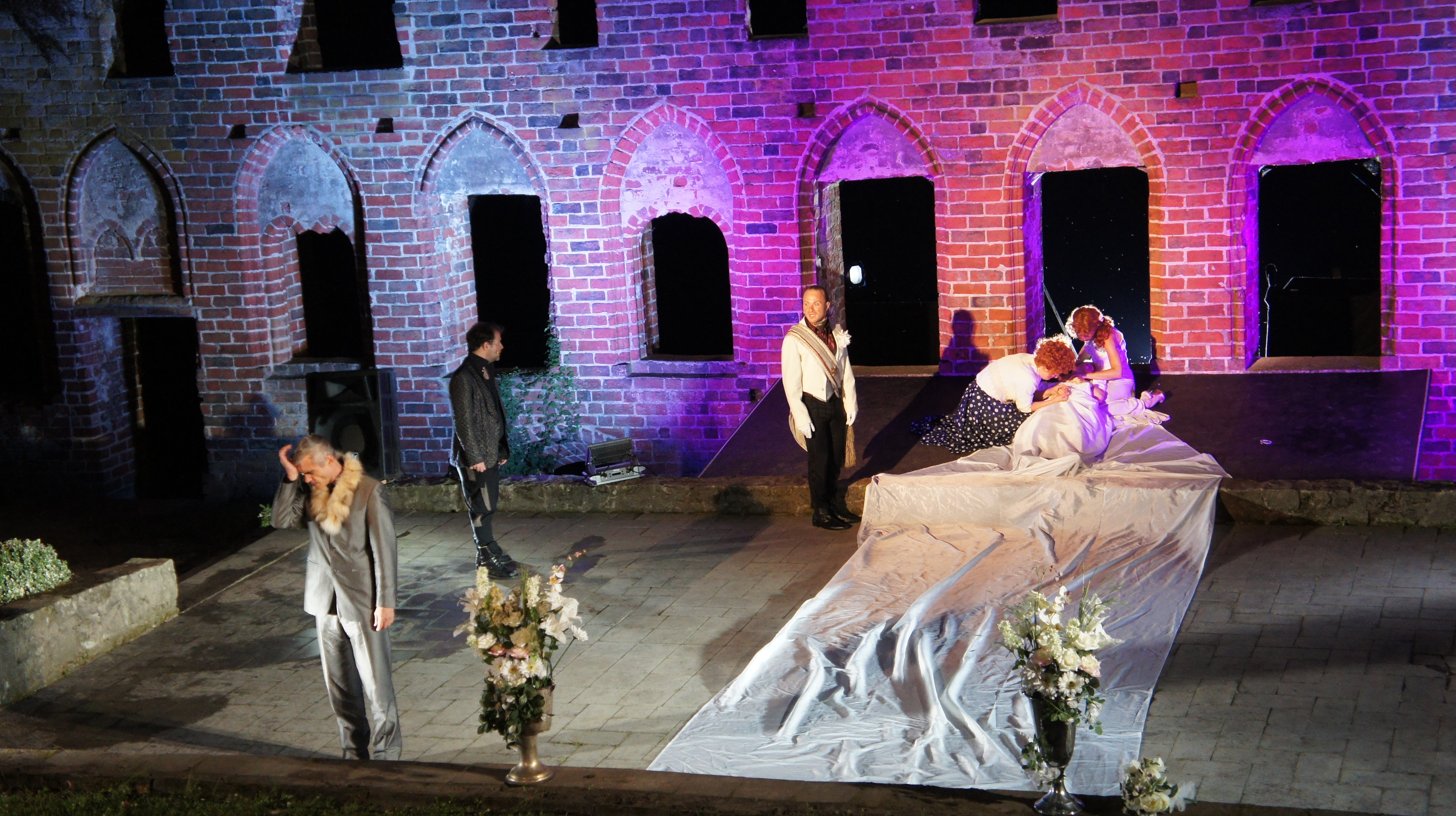 Theater im Kloster Arendsee. Fünf Schauspielende stehen verteilt auf einer Bühne vor den bunt beleuchteten Backsteinmauern.