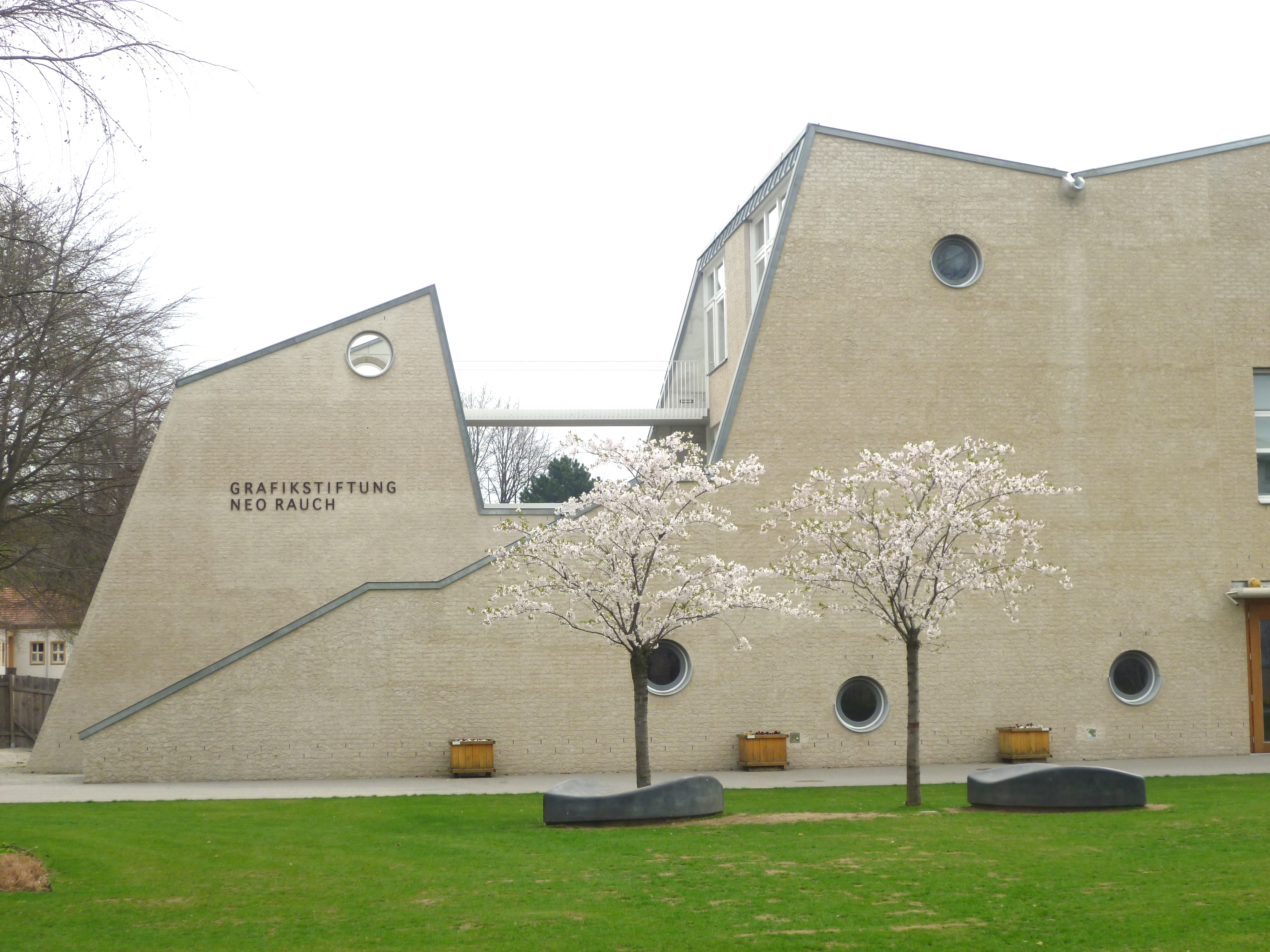 Die Außenansicht der Grafikstiftung Neo Rauch. Das Industriedenkmal mit seiner asymmetrischen Silhouette befindet sich in einem Park und ist umgeben von einer grünen Wiese und blühenden Bäumen.