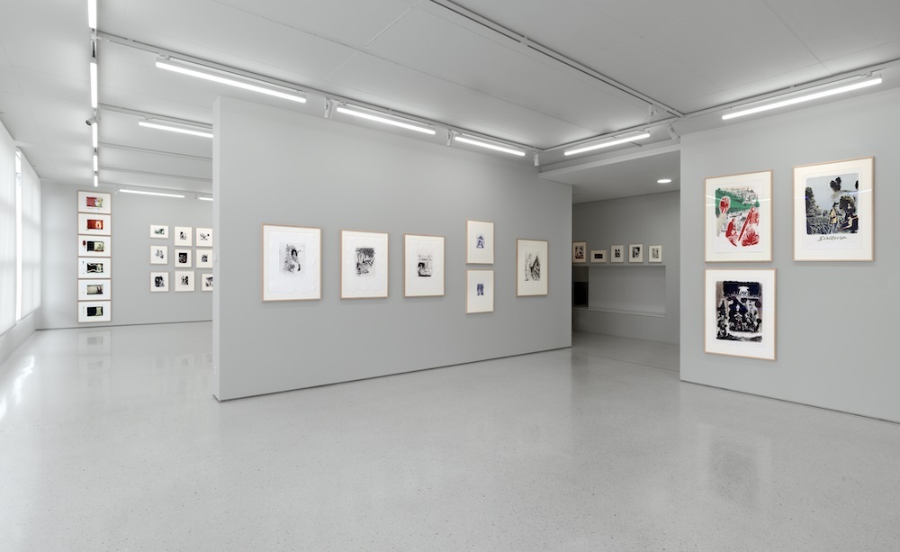 Blick in die Ausstellung "Neo Rauch Der Bestand Druckgrafik seit 1988". Druckgrafische Werke von Neo Rauch hängen an grauen Wänden. Helle Neonröhren beleuchten die Ausstellung.