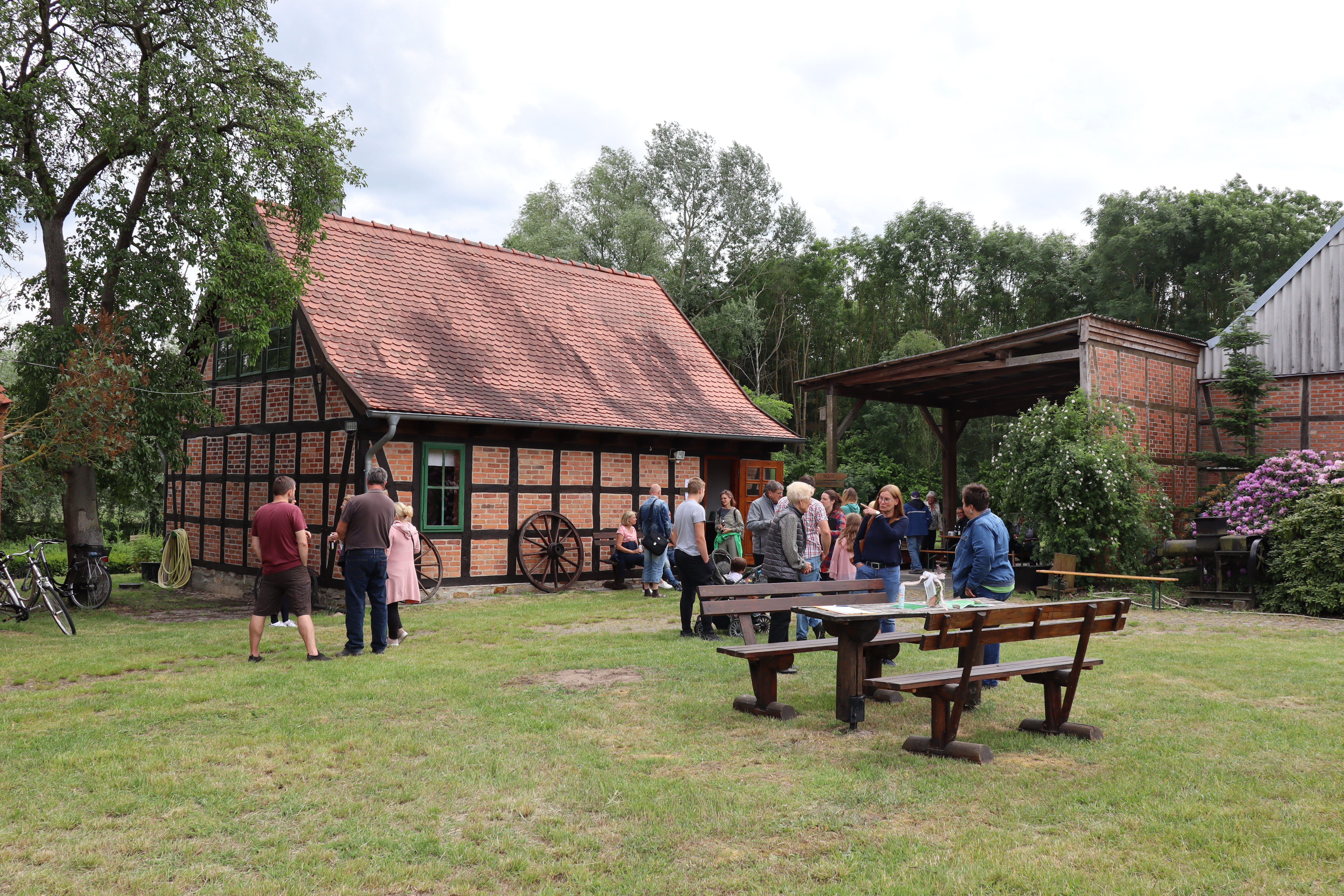 Zwei kleine Fachwerkhäuser aus Backstein stehen auf einer Wiese. Um sie herum steht eine Gruppe von Menschen.
