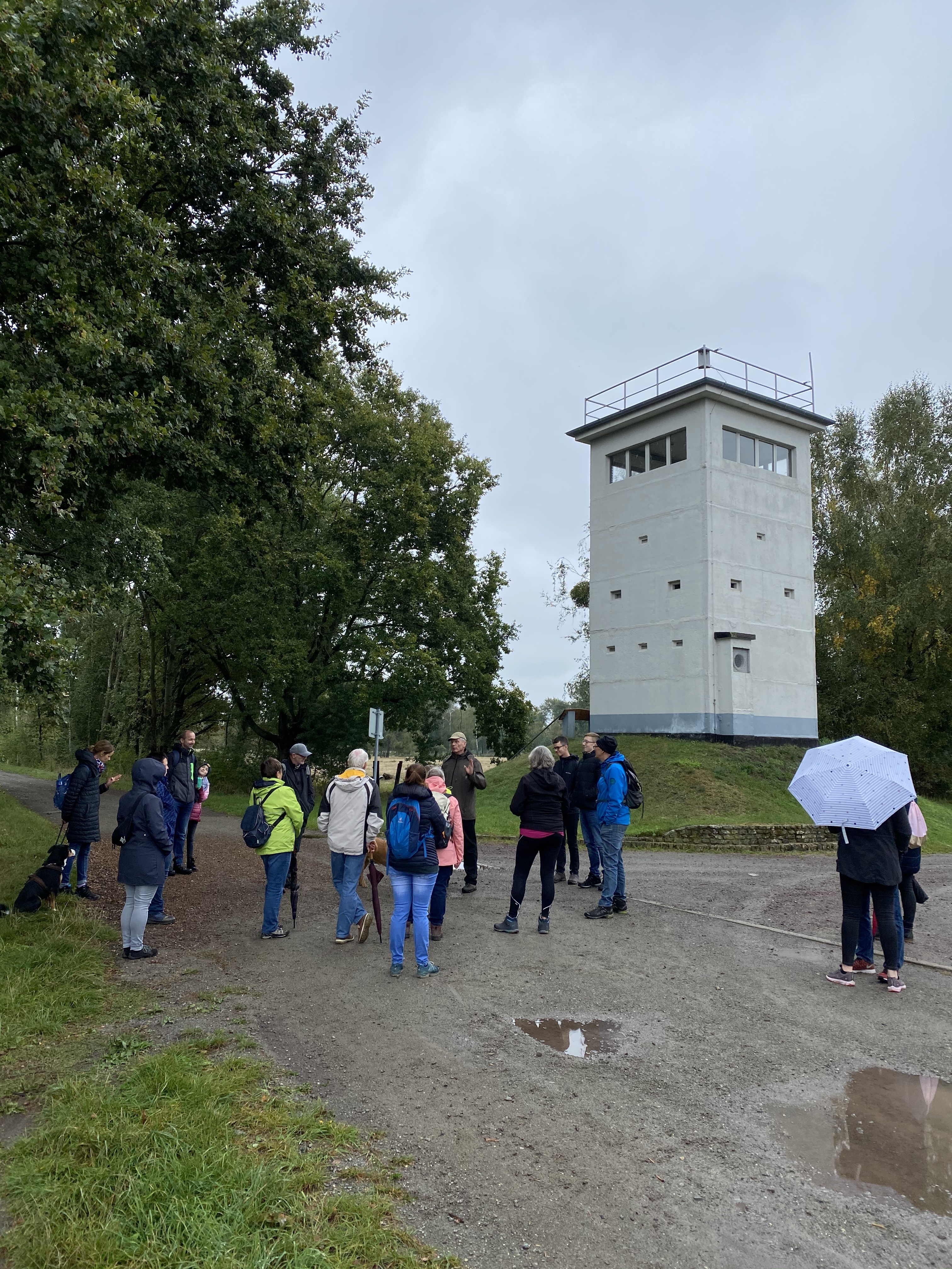 Ein niedriger grauer Grenzturm im Wald. Auf einem Weg steht eine Besuchergruppe davor.