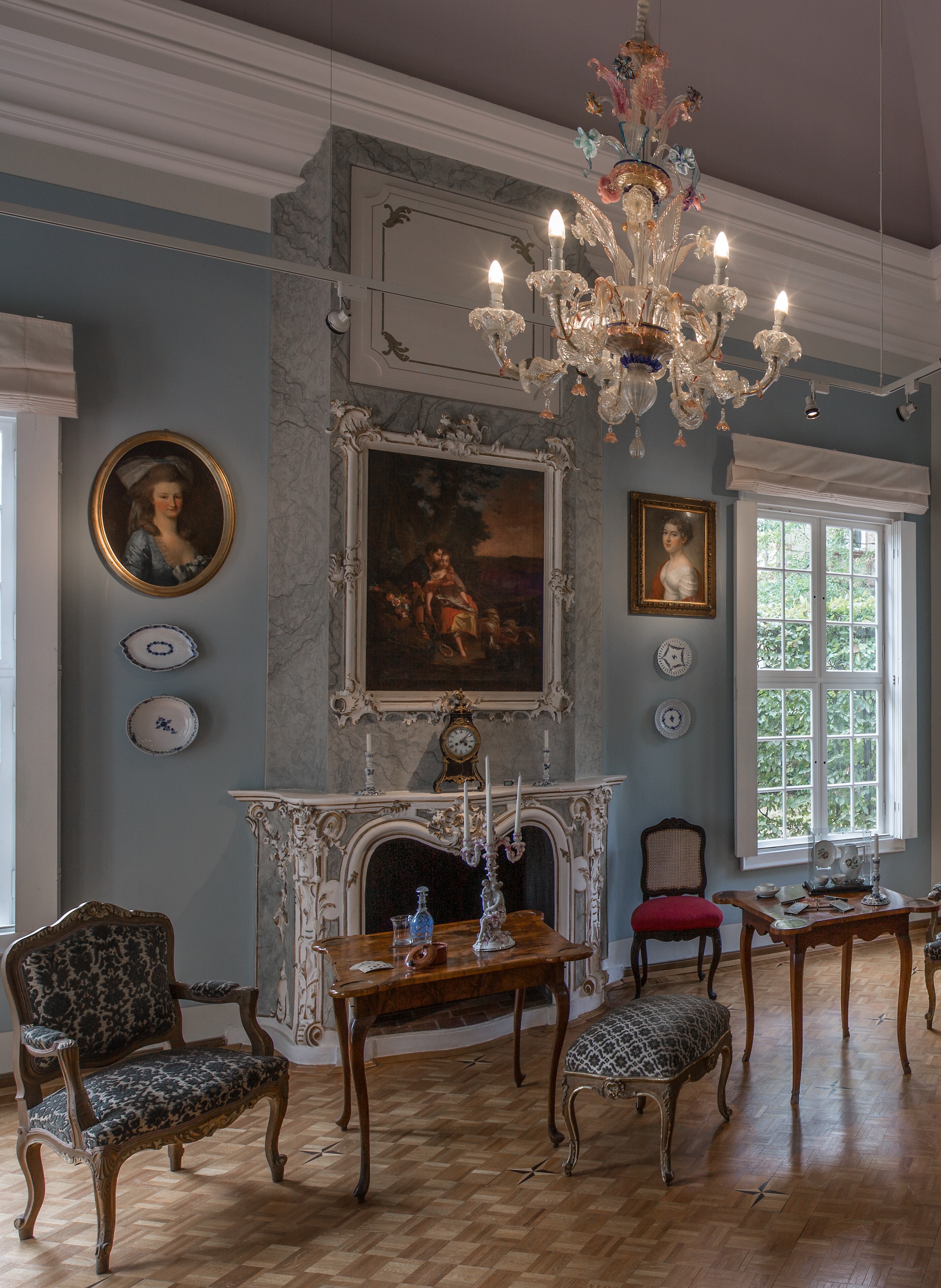 Innenraum des Herzogspavillon. Ein Spielsalon des 18. Jahrhunderts, mit gepolsterten Sitzgelegenheiten, einem Kamin und Porzellangeschirr an den Wänden und auf zwei Tischen stehend. Zwei Porträtgemälde zeigen zwei Frauen und rahmen ein größeres Gemälde. Ein Leuchter aus buntem Glas erhellt den Raum.
