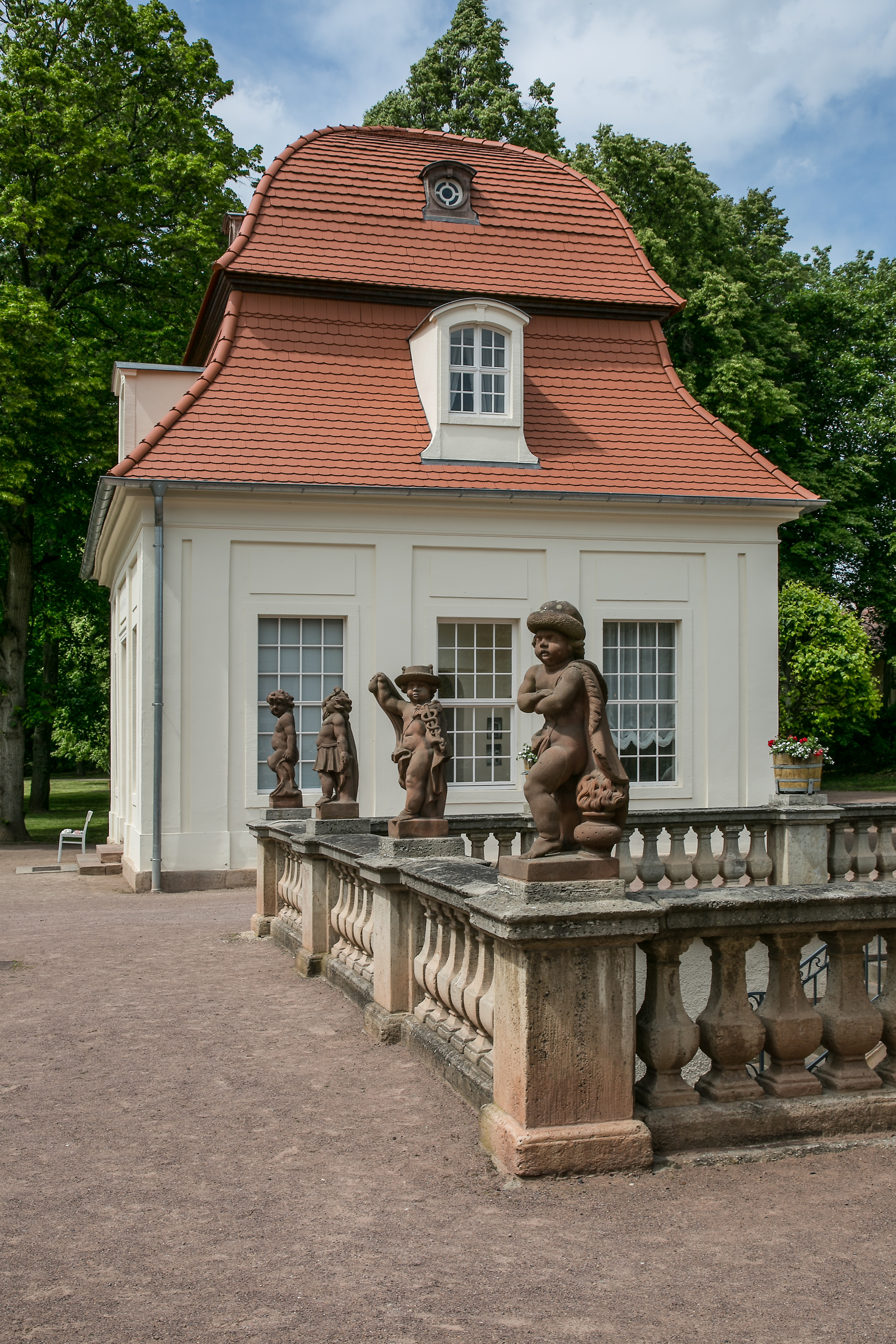 Brunnen der Balustrade der Historischen Kuranlagen Bad Lauchstädt. Auf der Balustrade stehen vier Putten verkleidet als antike Götter. Hinter ihnen steht ein weißer Pavillon mit einem Pagodendach.