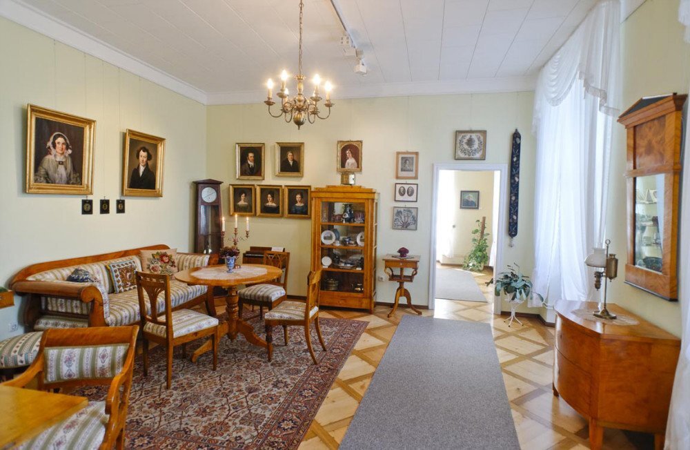 Im Kügelgenzimmer befindet sich eine Sitzgruppe aus mit Stoff bezogenen Holzmöbeln auf der linken Seite. Weitere Holzmöbel statten den Raum aus. Auf dem Boden liegt ein großer bunter Teppich. An den Wänden hängen Porträts.