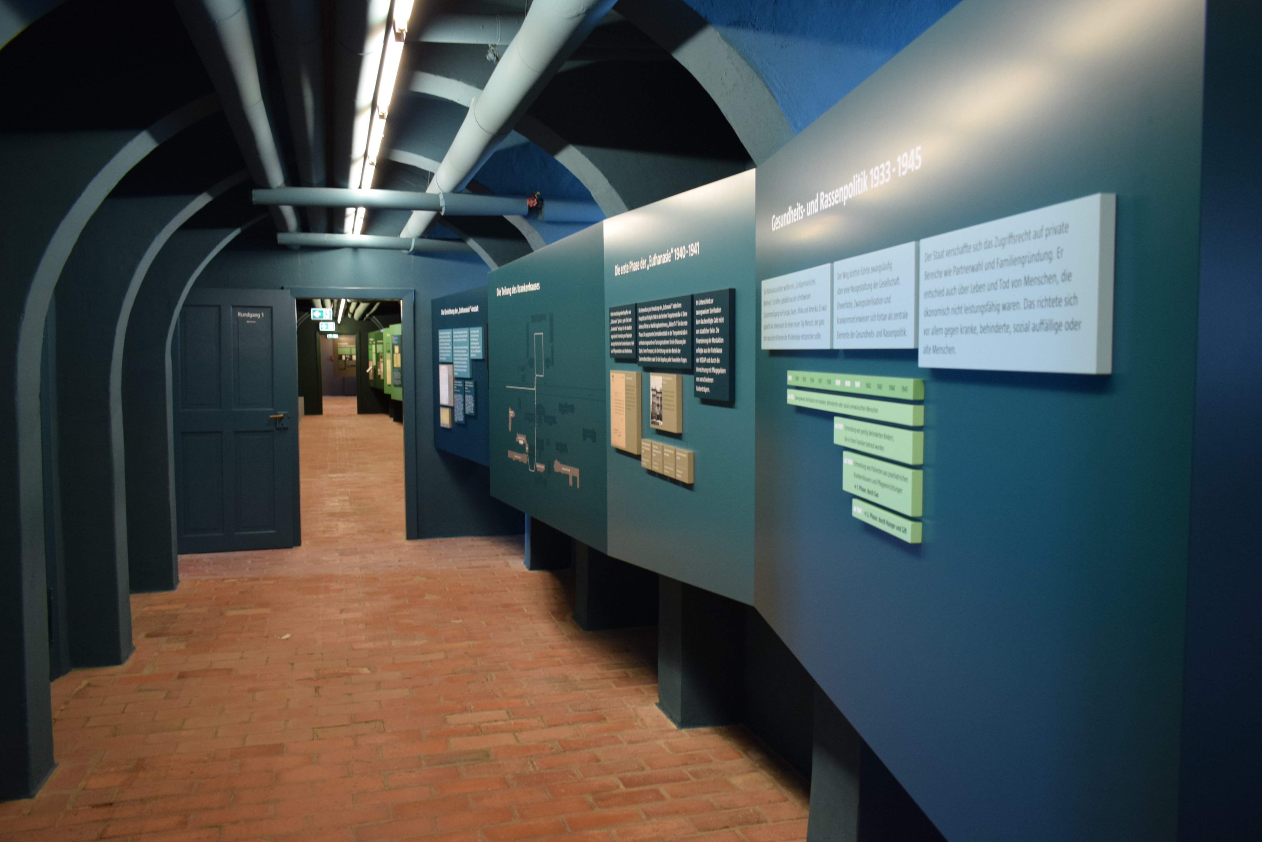Ein tunnelähnlicher blauer Gang führt durch die Dauerausstellung. An der rechten Seite befinden sich Informationstafeln.