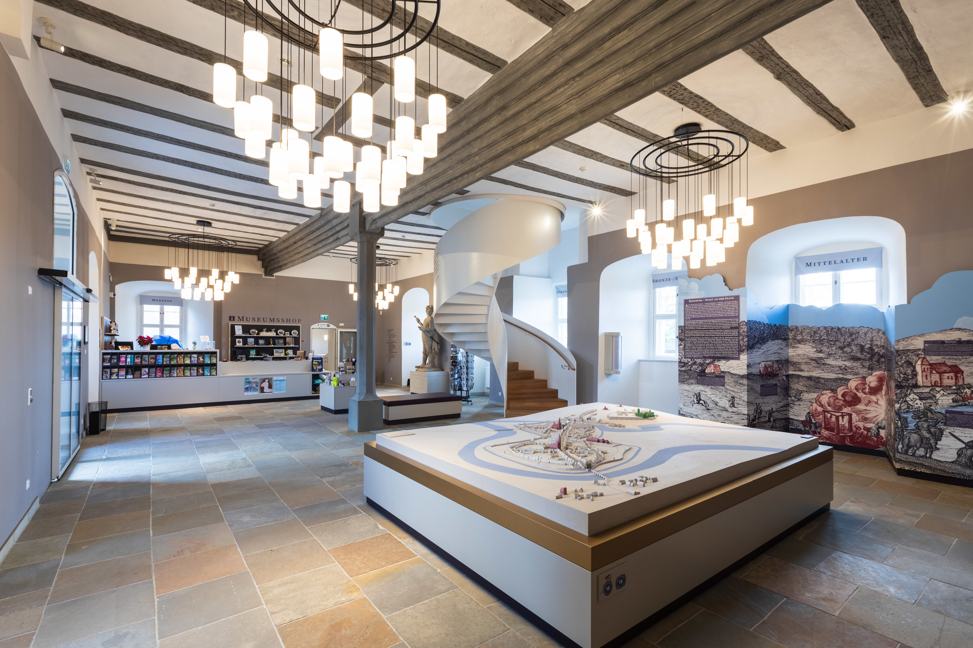 Hofstube im Museum Schloss Bernburg. Eingangsbereich mit einem Stadtmodell in der Mitte des Raumes. Aufsteller an der Seite informieren über das Mittelalter. Im Hintergrund führt eine weiße Wendeltreppe in die Ausstellung.