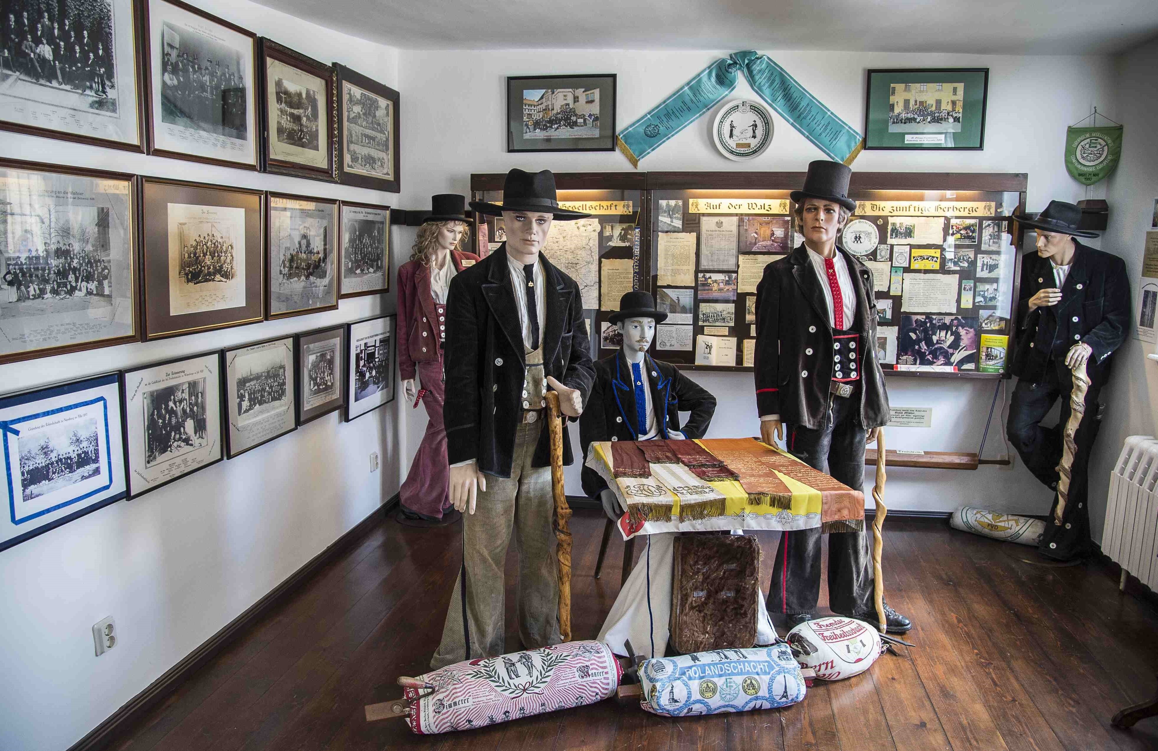 Ausstellungsraum "Die Walz" Darstellung der reisenden Schächte. Vier Modelle präsentieren die Kleidung der Gesellen: sie tragen einen schwarzen Hut, ein schwarzes Jackett oder eine Weste mit acht Knöpfen und eine Hose mit Schlag. Eine weibliche Puppe trägt die Kleidung in der Farbe rot. An den wänden hängen historische Gruppenbilder der Gesellen.