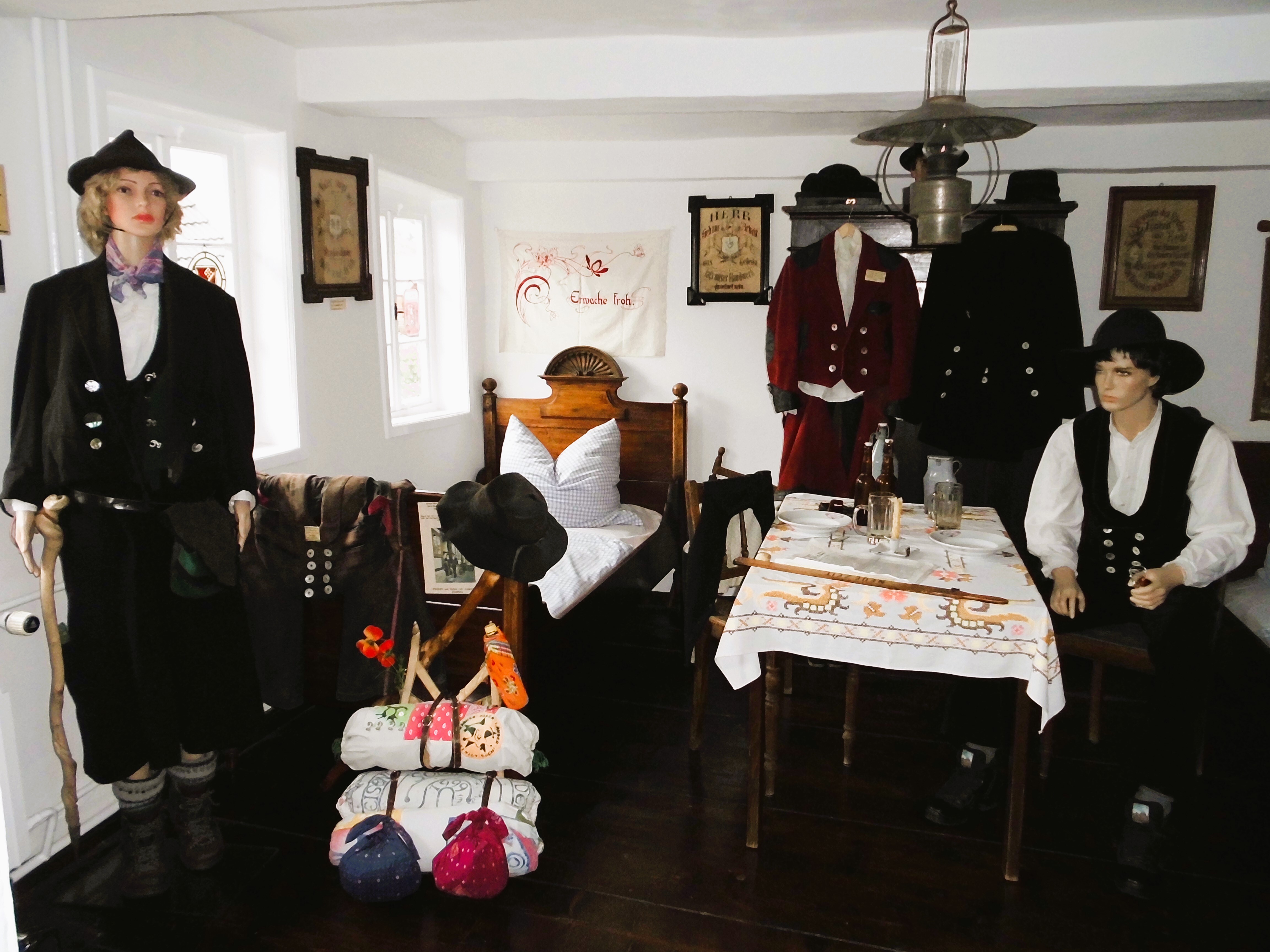 Logierzimmer (Schlafzimmer zu Zeiten der Herberge). Ein kleiner Raum mit zwei Betten. An einem Tisch in der Mitte des Raumes sitzt ein Geselle. Hinter ihm hängt die rote und schwarze Gesellenkleidung. Ein weiterer Geselle steht vor einem der Betten mit einem gepackten Rucksack.