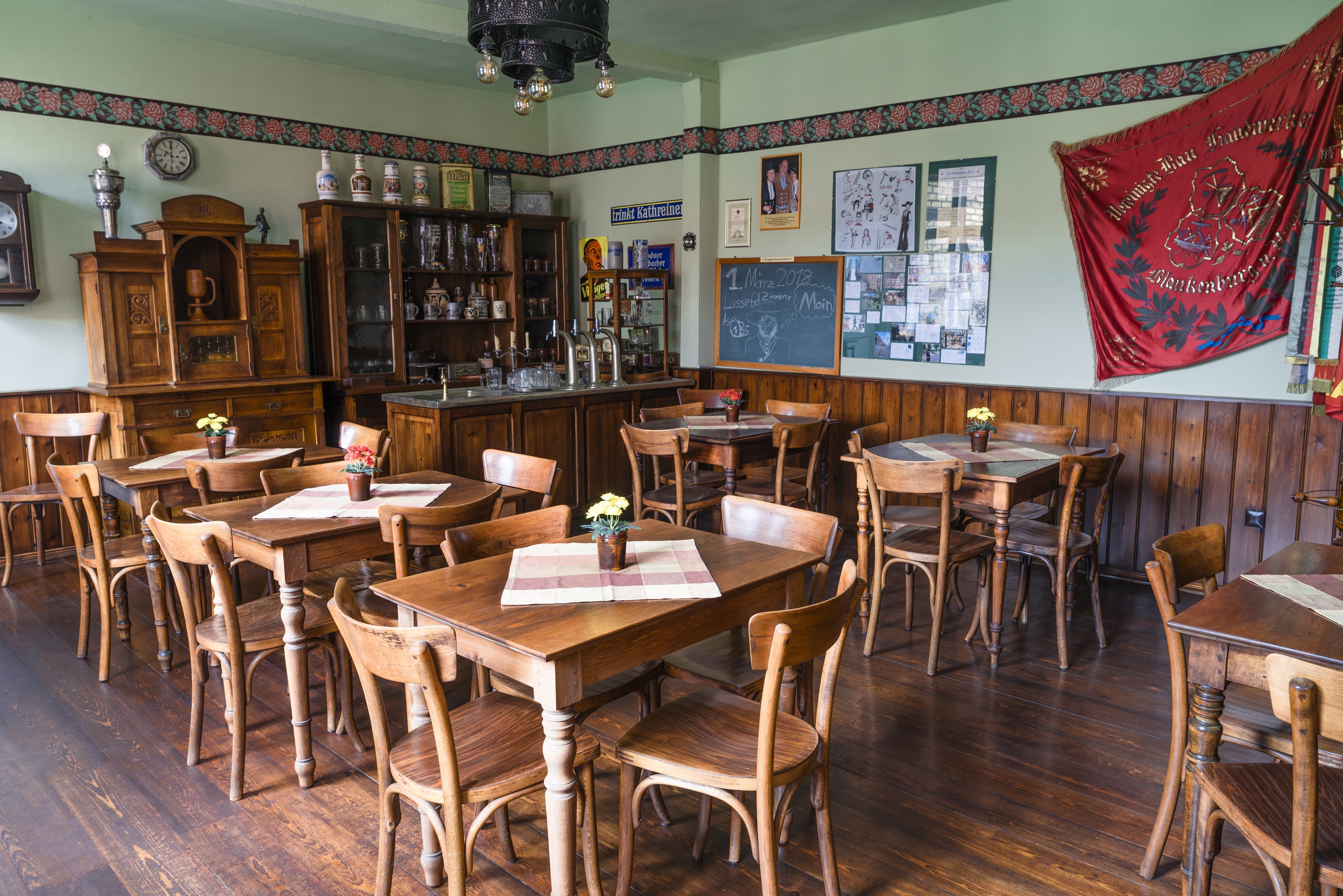 Herbergsmuseum - Restaurationzimmer (Herbergskneipe). Eine Kneipe mit Tischen und Stühlen aus Holz. An der Wand hängen eine große rote Fahne und Bilder von Gesellen.