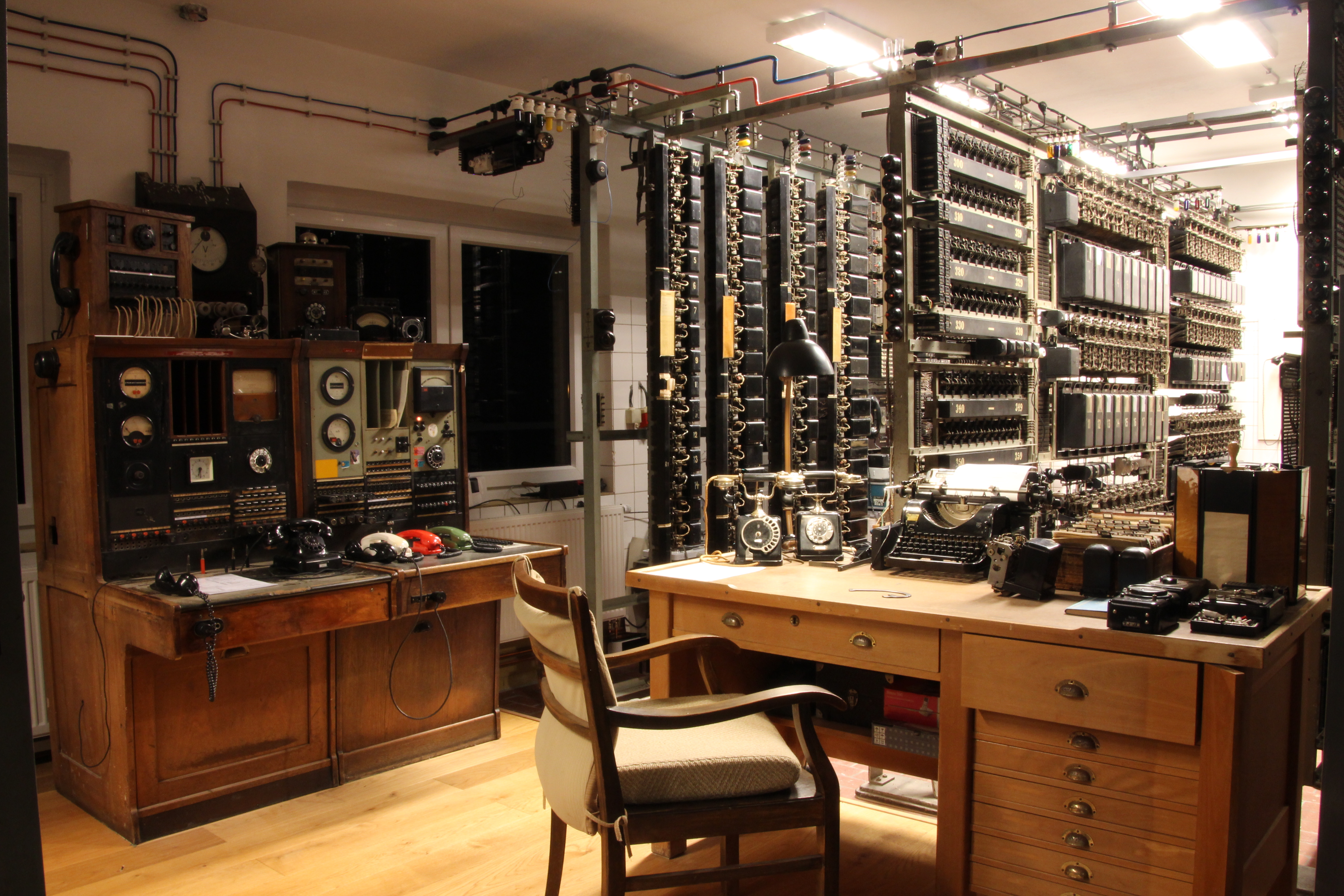 Im Wählersaal steht ein Schreibtisch mit einer Schreibmaschine, zwei Telefonapparaten und verschiedenen Kameras. In einem Schrank daneben sind drei bunte Telefonhörer angeschlossen. Mehrere Regalreihen beinhalten weitere technische Geräte.