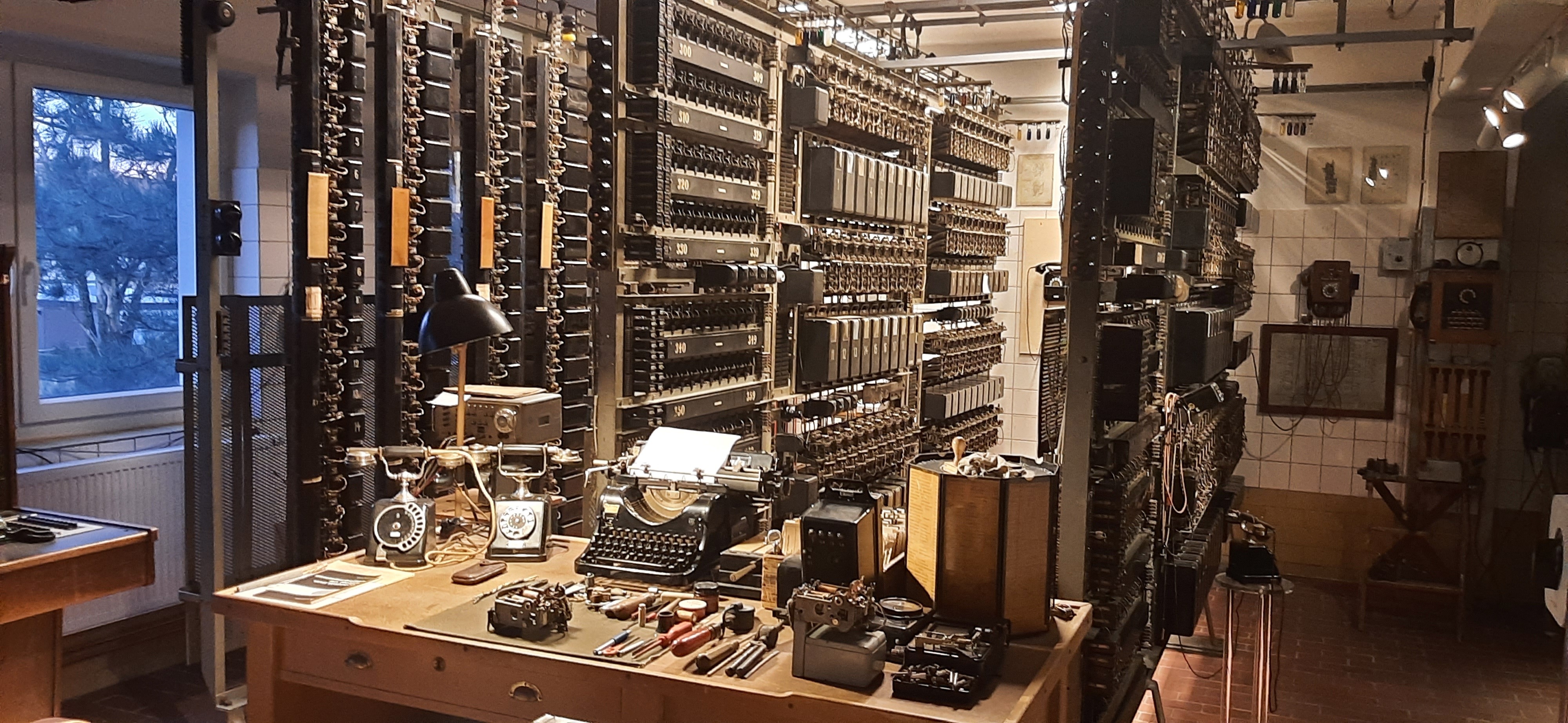 Im Saal steht ein großer Holzschreibtisch auf dem eine Schreibmaschine, zwei alte Telefone, Kameras und Werkzeuge stehen. Dahinter befinden sich mehrere Regale mit weiteren technischen Apparaturen.