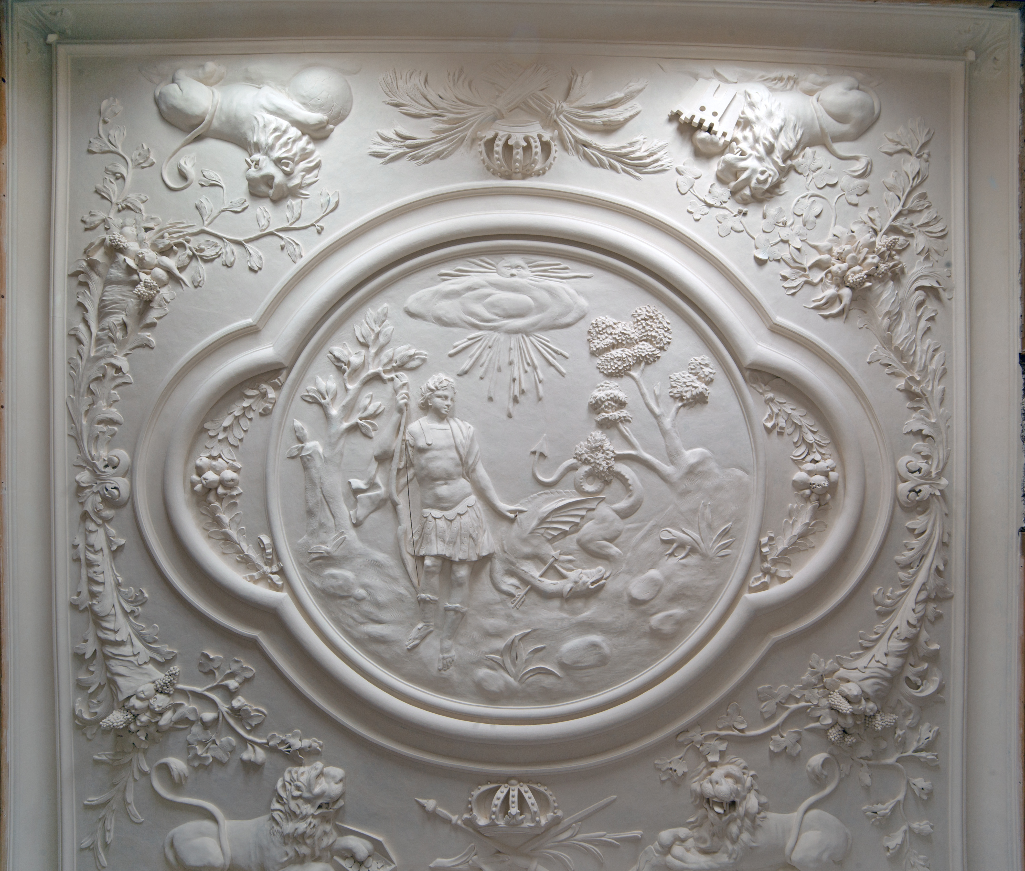 Stuckdecke mit Motiven aus der Antike. Eine weiße Stuckdecke mit einem großen Medaillon, in dem ein junger Mann mit einem Bogen steht in der Landschaft. Ein kleiner Drache liegt neben ihm, ein Pfeil steckt in seinem Hals. Außerhalb der Szene befindet sich in den vier Ecken jeweils ein Löwe.