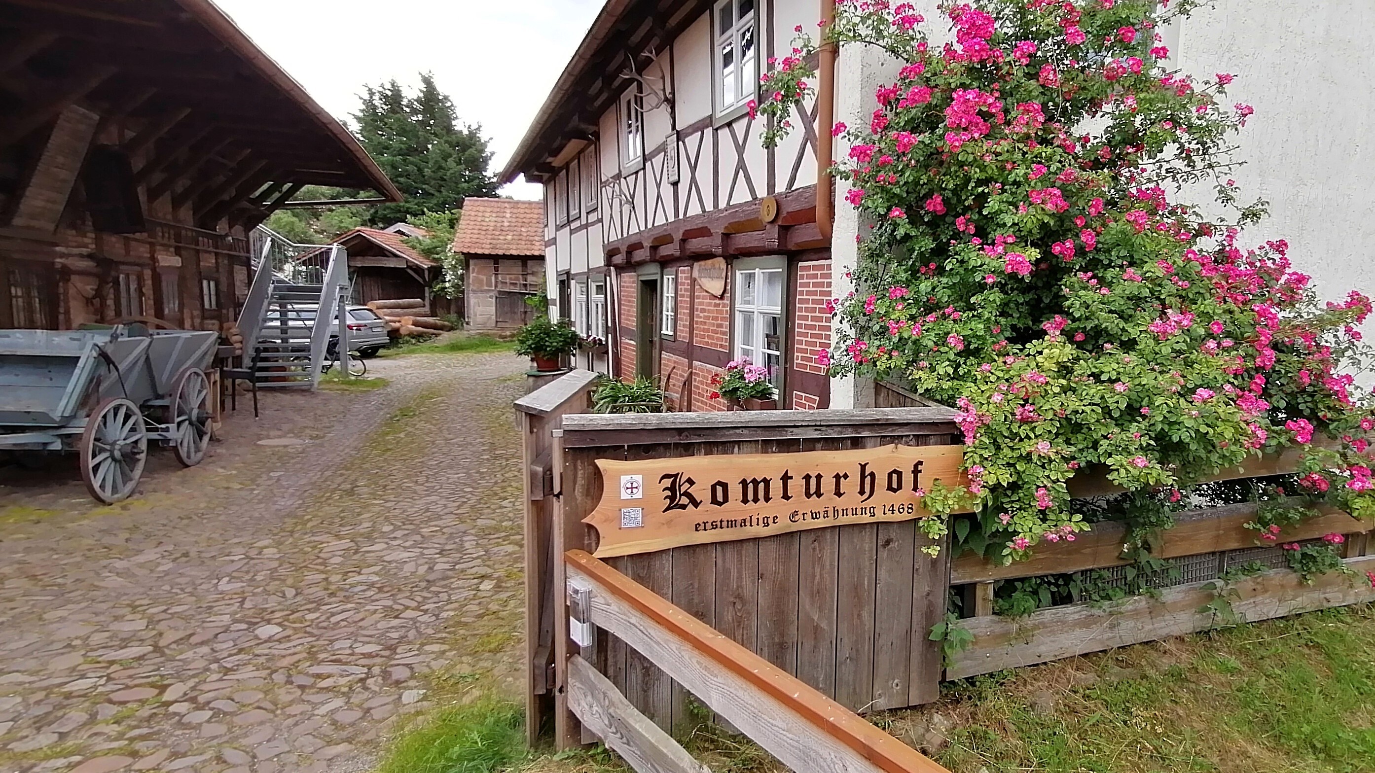 Der Komturhof Darlingerode. auf der rechten Seite befindet sich ein Fachwerkhaus. Gegenüber steht eine Scheune aus Holz mit einem blauen Holzwagen davor. Im Hintergrund sind zwei kleine Ställe erkennbar.