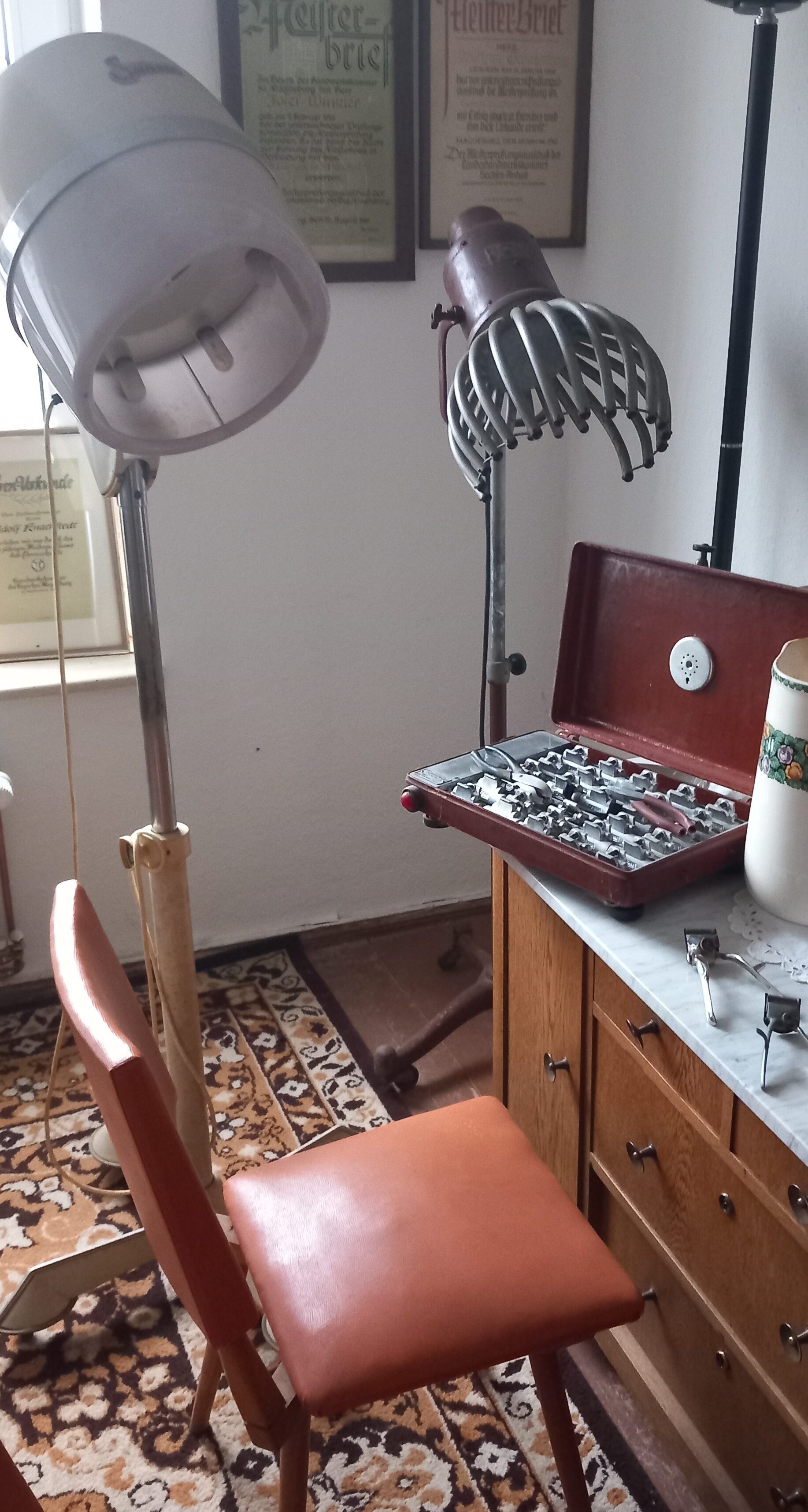 Neben einem Stuhl mit einem orangenen Bezug stehen eine alte Trocken- und Fingerhaube. Auf einem Schrank steht weiteres Friseurzubehör.