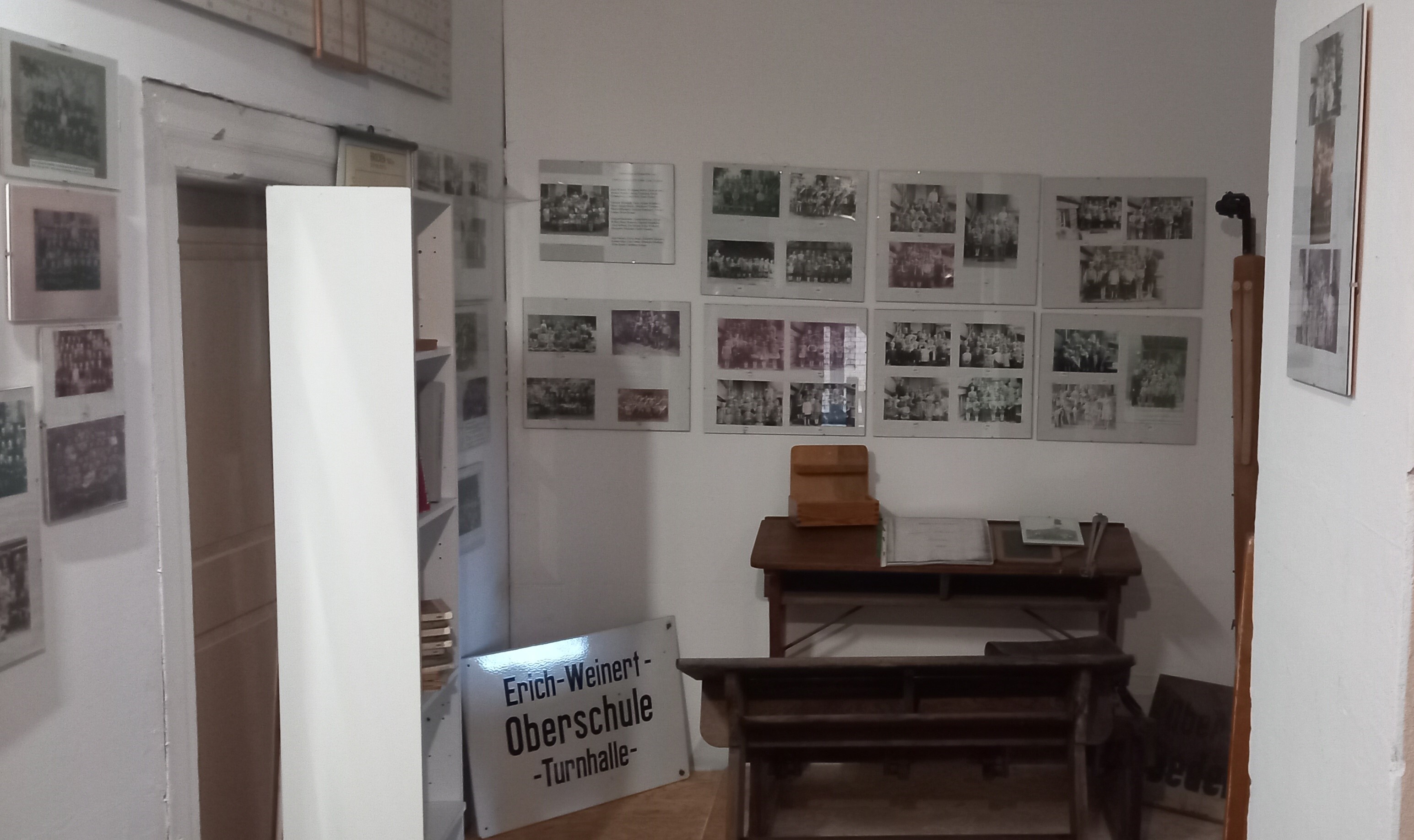 Eine Schulbank aus dunkelbraunem Holz steht an einer Wand. An den Wänden hängen Fotografien in schwarz-weiß. Vor einer Tür steht ein weißes Regal mit Büchern.