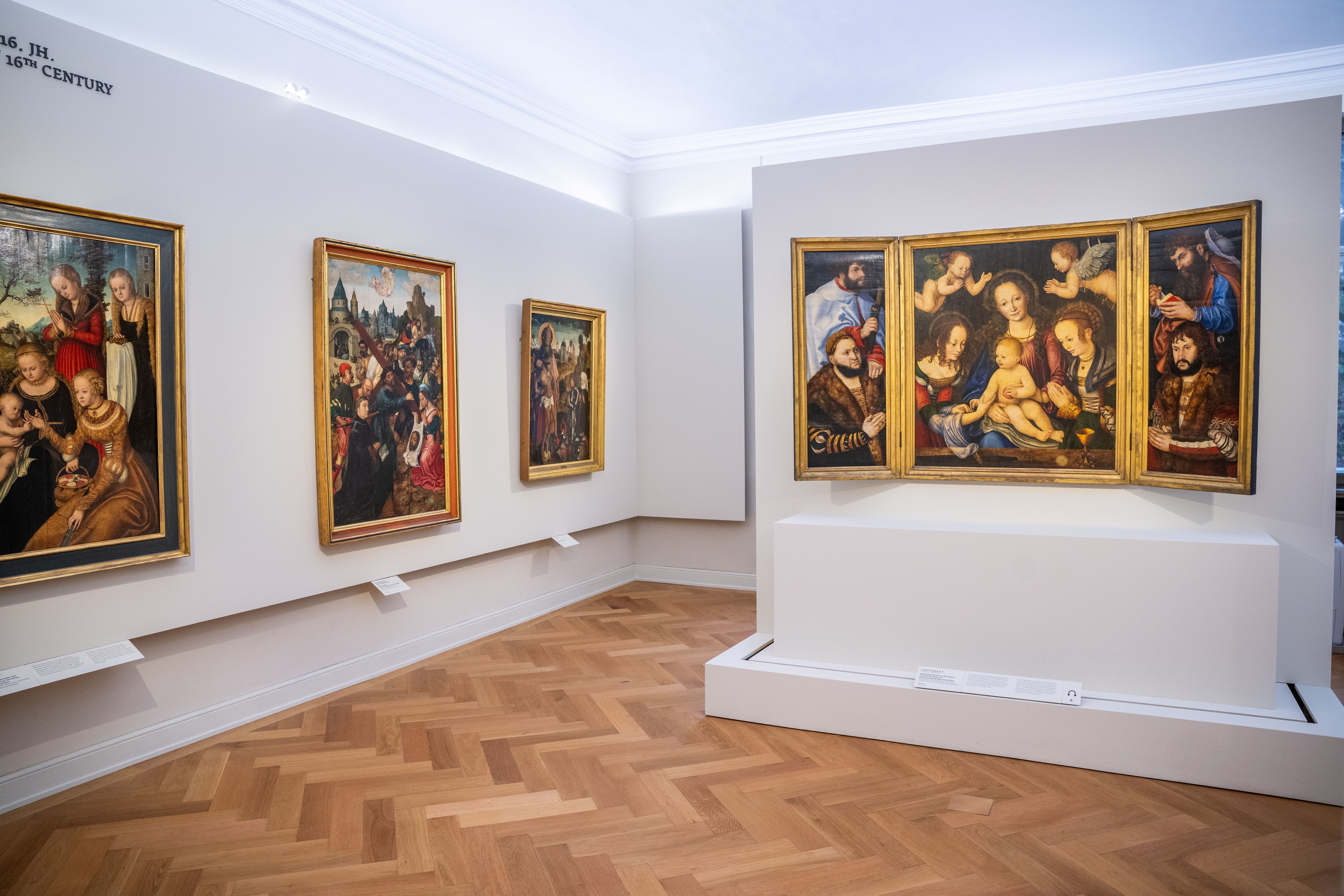 In einem weißen Ausstellungsraum hängen Gemälde und ein Altar von Lucas Cranach.