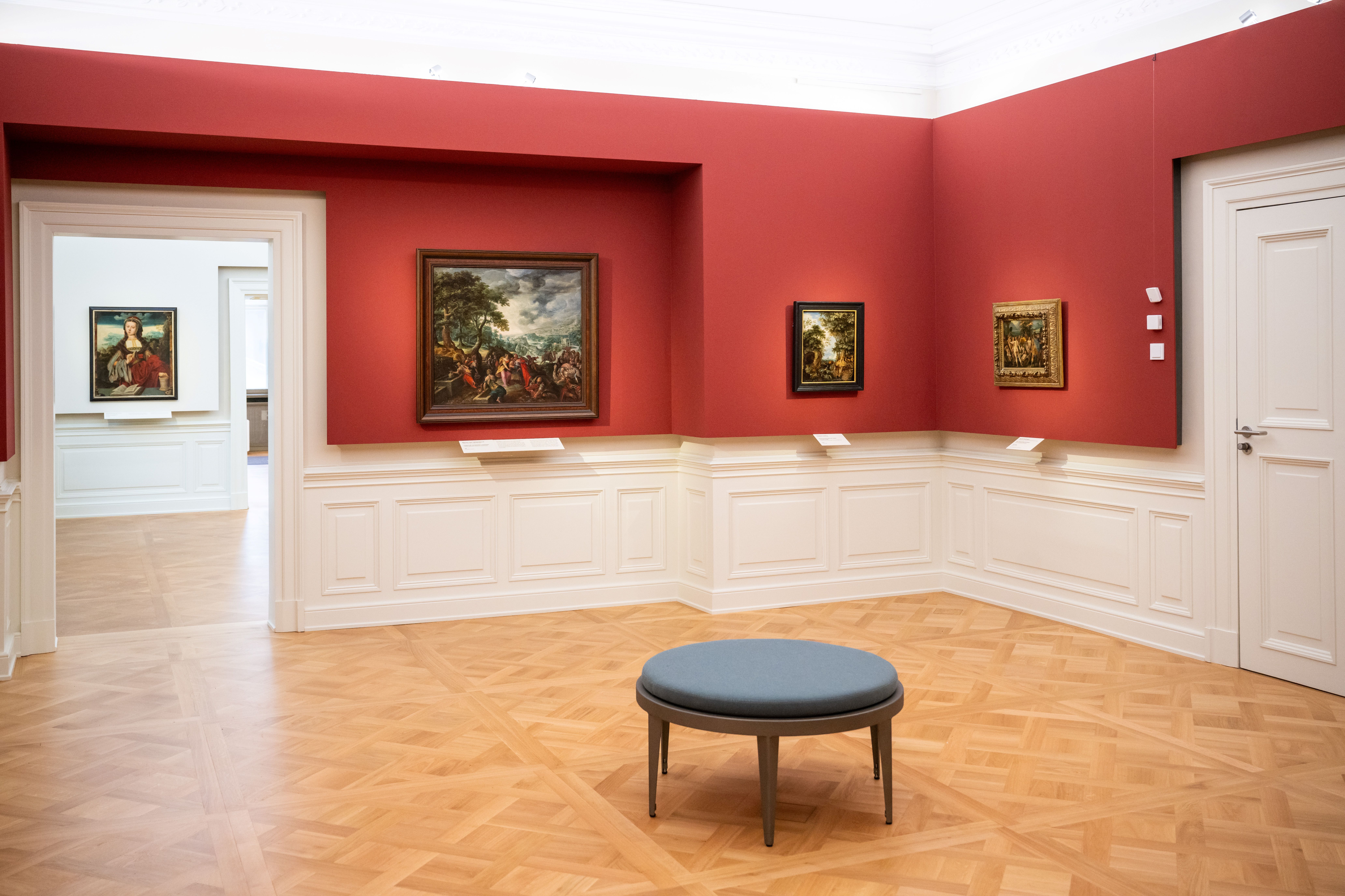 Ein roter Ausstellungsraum mit einem Hocker in der Mitte. An den Wänden hängen Gemälde.