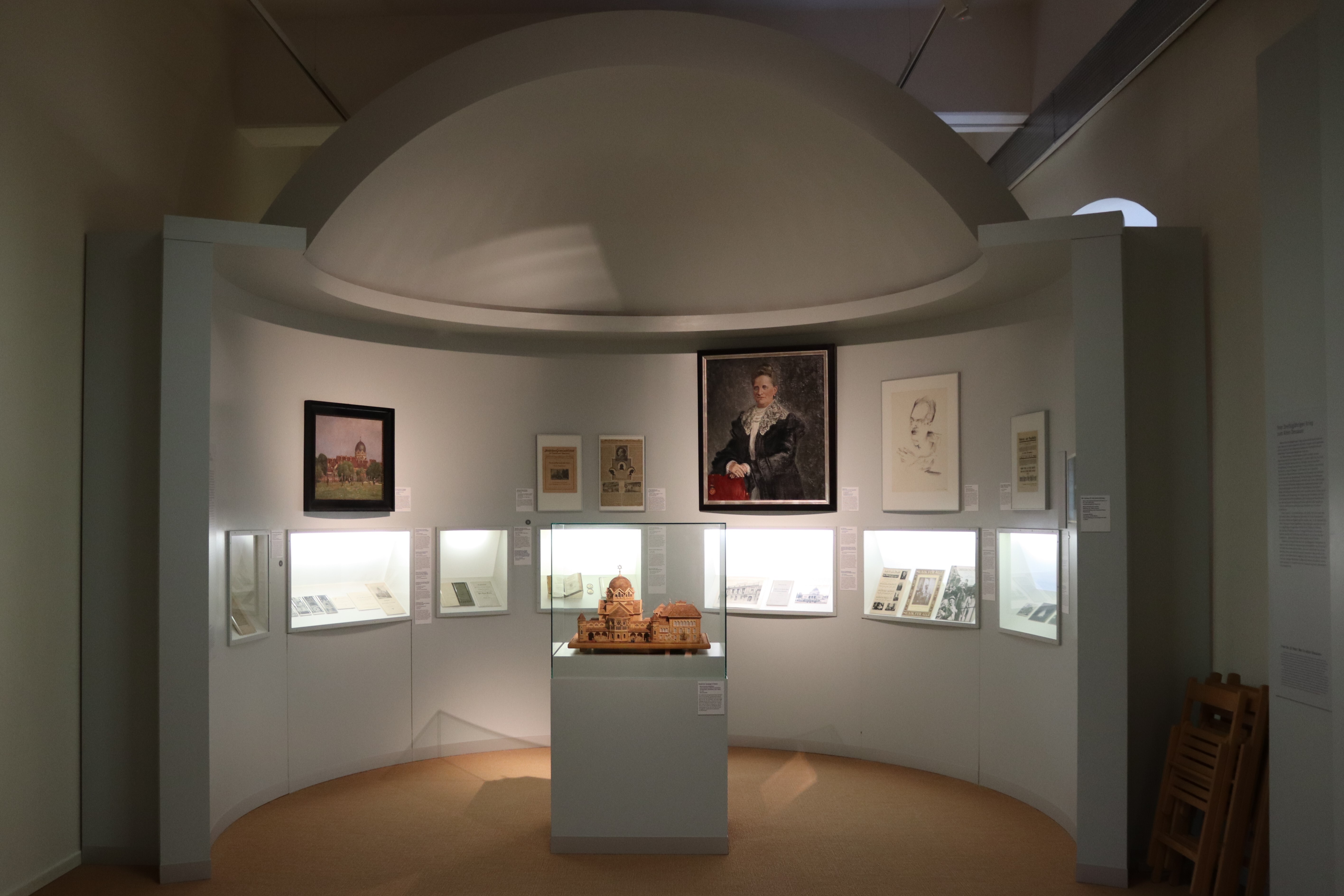 In einer weißen halbkreisförmigen Kalotte steht das Modell eines Gebäudes. An den Wänden hängen Gemälde und es sind kleine Vitrinen darin eingelassen.