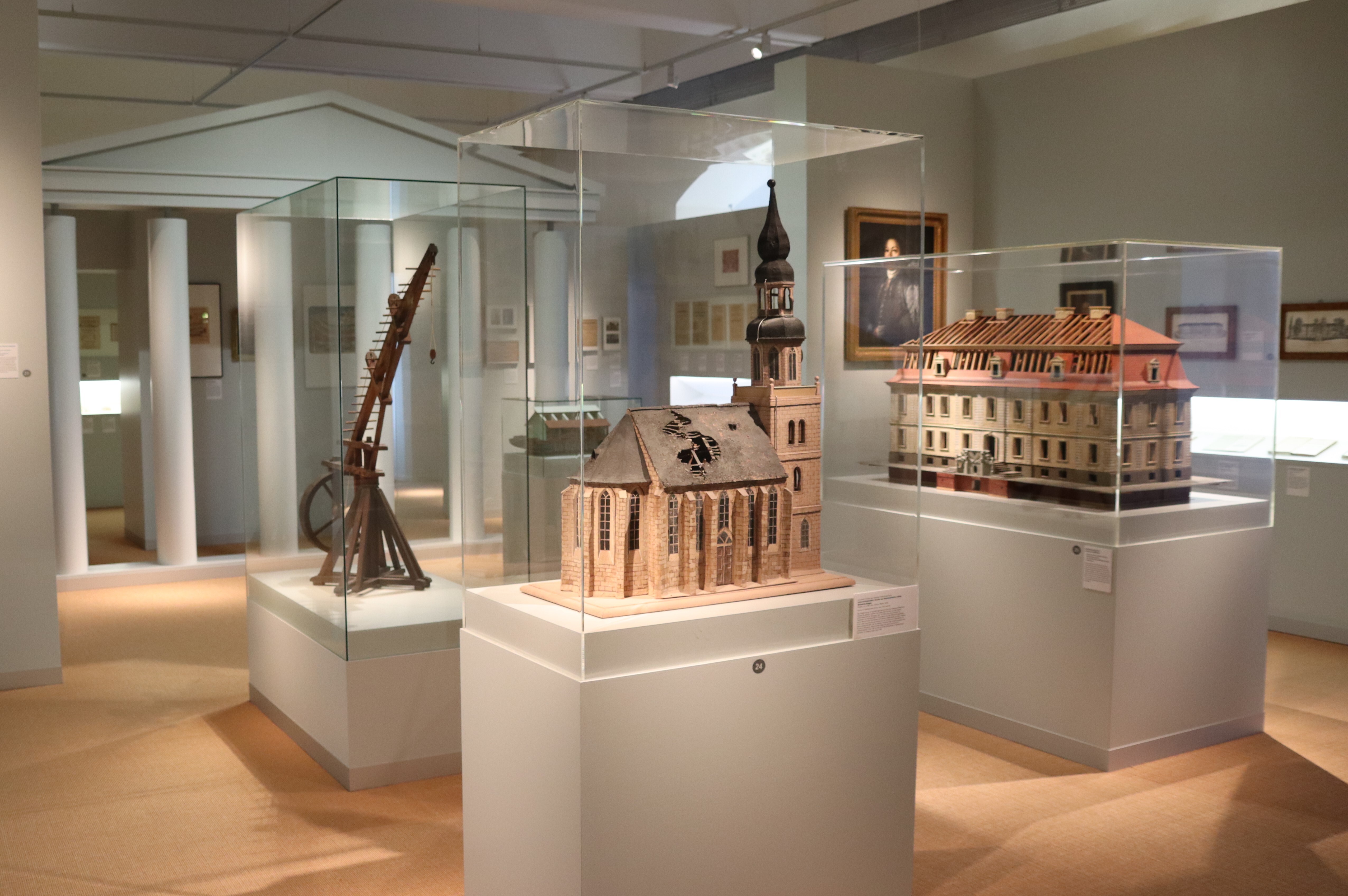 Blick in die Ausstellung. Drei Architekturmodelle stehen in Vitrinen im Raum. Ein weiteres Modell eines technischen Gerätes aus Holz befindet sich in einer weiteren Vitrine.