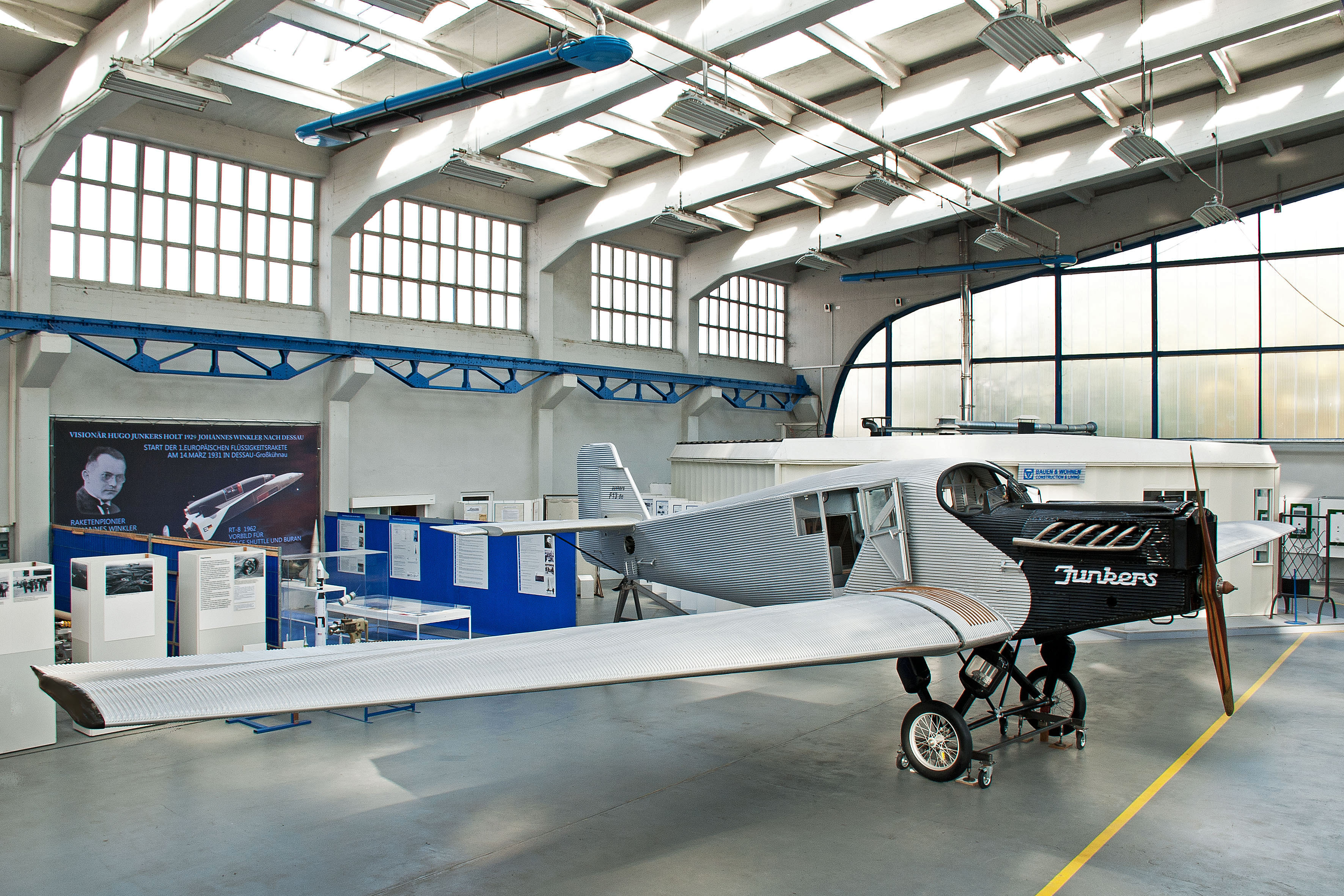 Junkers F13. In einer hellen Lagerhalle steht ein kleines Flugzeug des Modells "Junkers F13". Es ist weiß und hat eine schwarze Flugzeugnase. Dahinter ist ein Teil der Ausstellung zu sehen: mehrere Vitrinen mit Aufstellwänden und ein großes Plakat sind zu sehen.