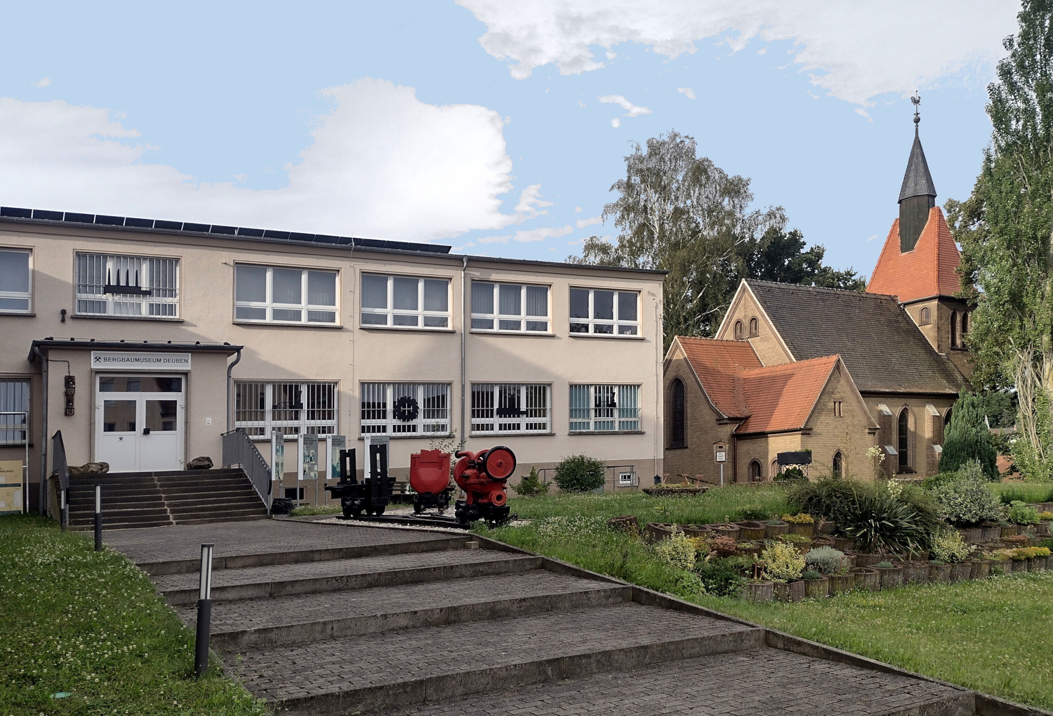 Bergbaumuseum Deuben Außenansicht. Ein heller Flachbau mit der Bezeichnung "Bergbaumuseum Deuben" steht neben einer kleinen Kirche. Vor dem Museum stehen rote Bergbaumaschinen.