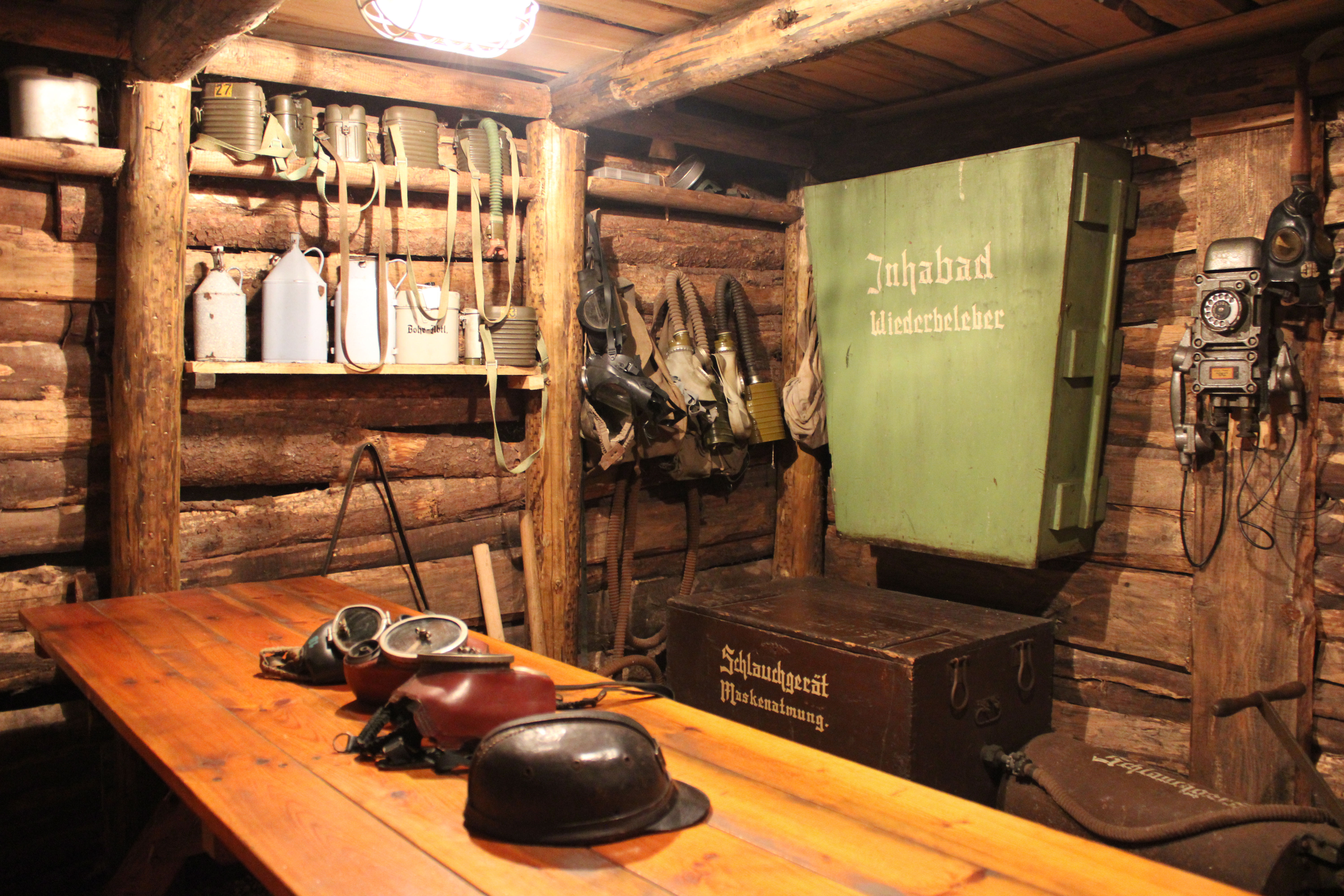 Auf einem Holztisch liegen ein Helm und Atemmasken der Bergarbeiter. Verschiedene Flaschen und Ferngläser stehen in einem Regal an einer Holzwand. Ein großer grüner Kasten, eine braune Kiste und ein altes graues Telefon befinden sich an der Wand daneben.
