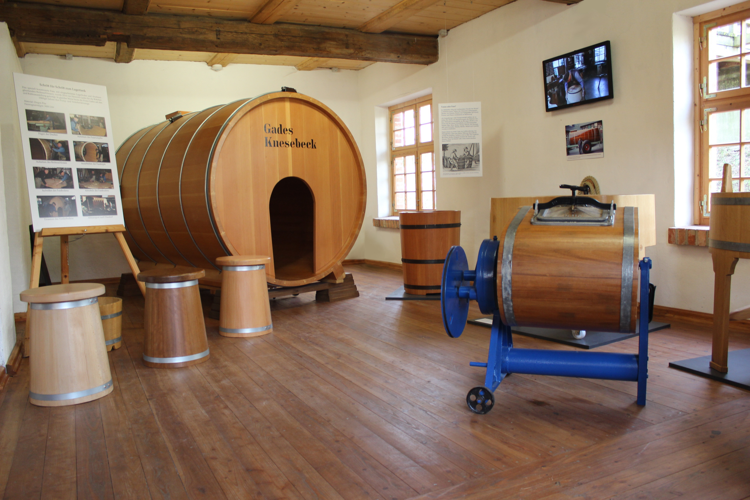Freilichtmuseum Diesdorf Böttcherei und Küferei Ausstellung. Ein großes Holzfass mit einer Öffnung steht in einem Raum. Weitere kleinere Fässer stehen im Raum verteilt. Eine Tafel mit Bildern und ein Film erklären die Herstellung der Fässer.