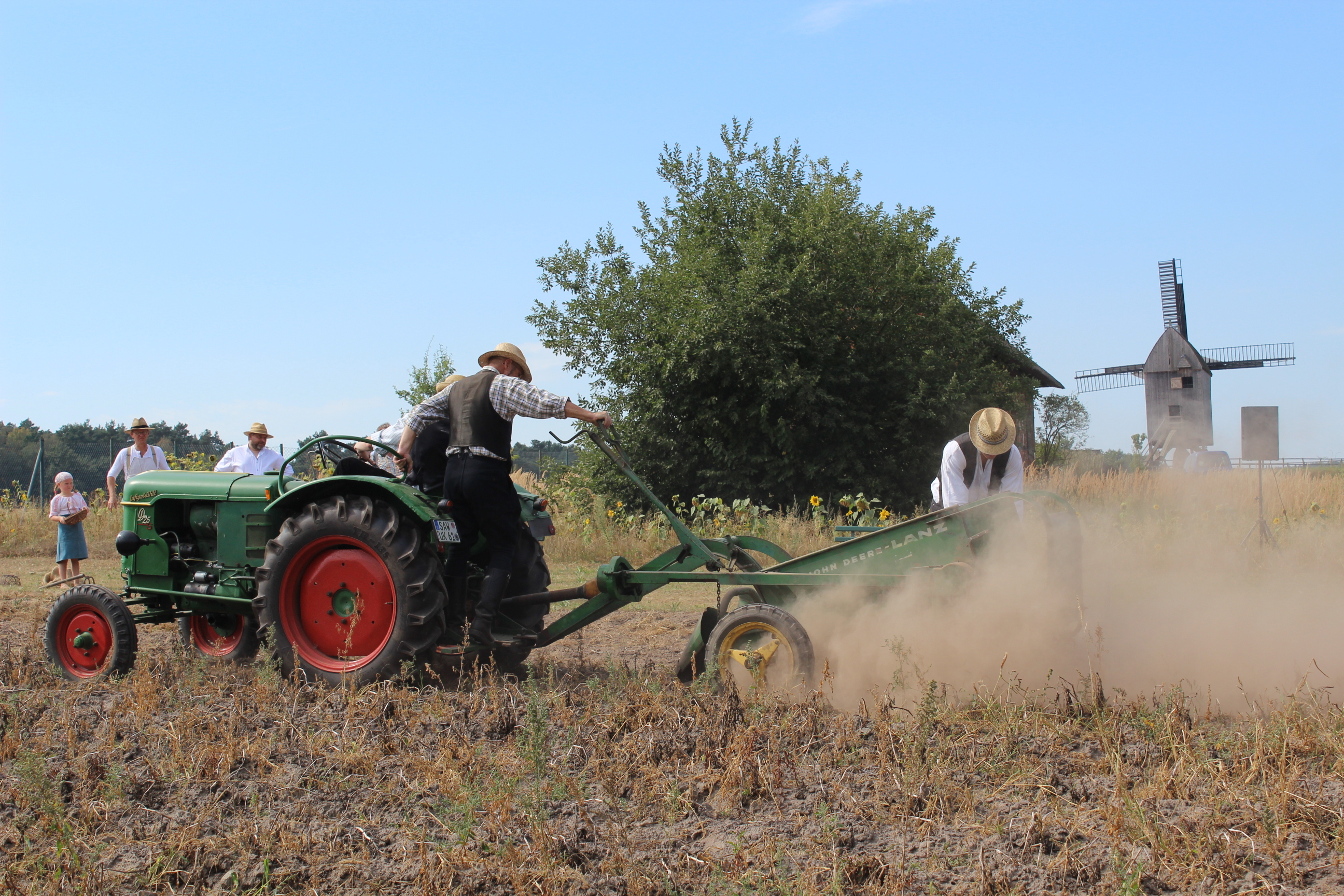 Freilichtmuseum Diesdorf: Kartoffelernte. Zwei Männer sitzen auf einer Ackermaschine, die über das Feld fährt. Drei weitere Männer und ein junges Mädchenschauen zu oder helfen bei der Arbeit. Eine kleine Windmühle steht im Hintergrund.