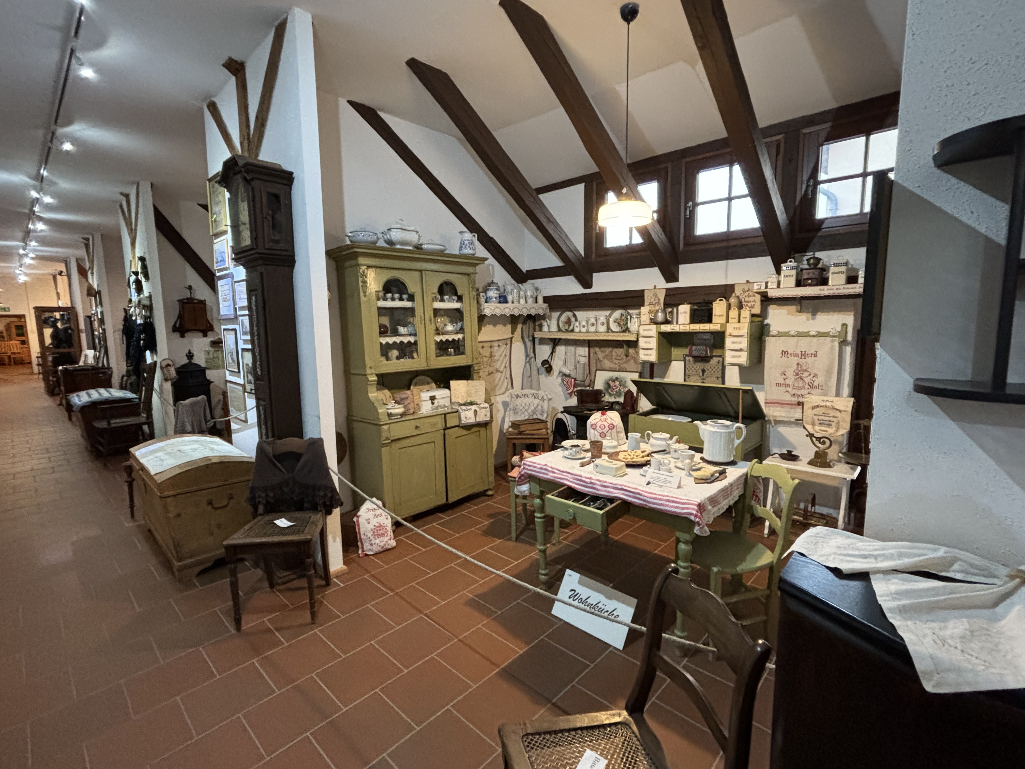 Heimatmuseum Ditfurt. Entlang eines Ganges sind kleine Wohnräume eingerichtet. Im ersten Raum ist eine Wohnküche ausgestellt. Die Küchenschränke und der Esstisch mit zwei Stühlen sind grün gestrichen. Überall stehen Haushaltsgeräte und Geschirr.