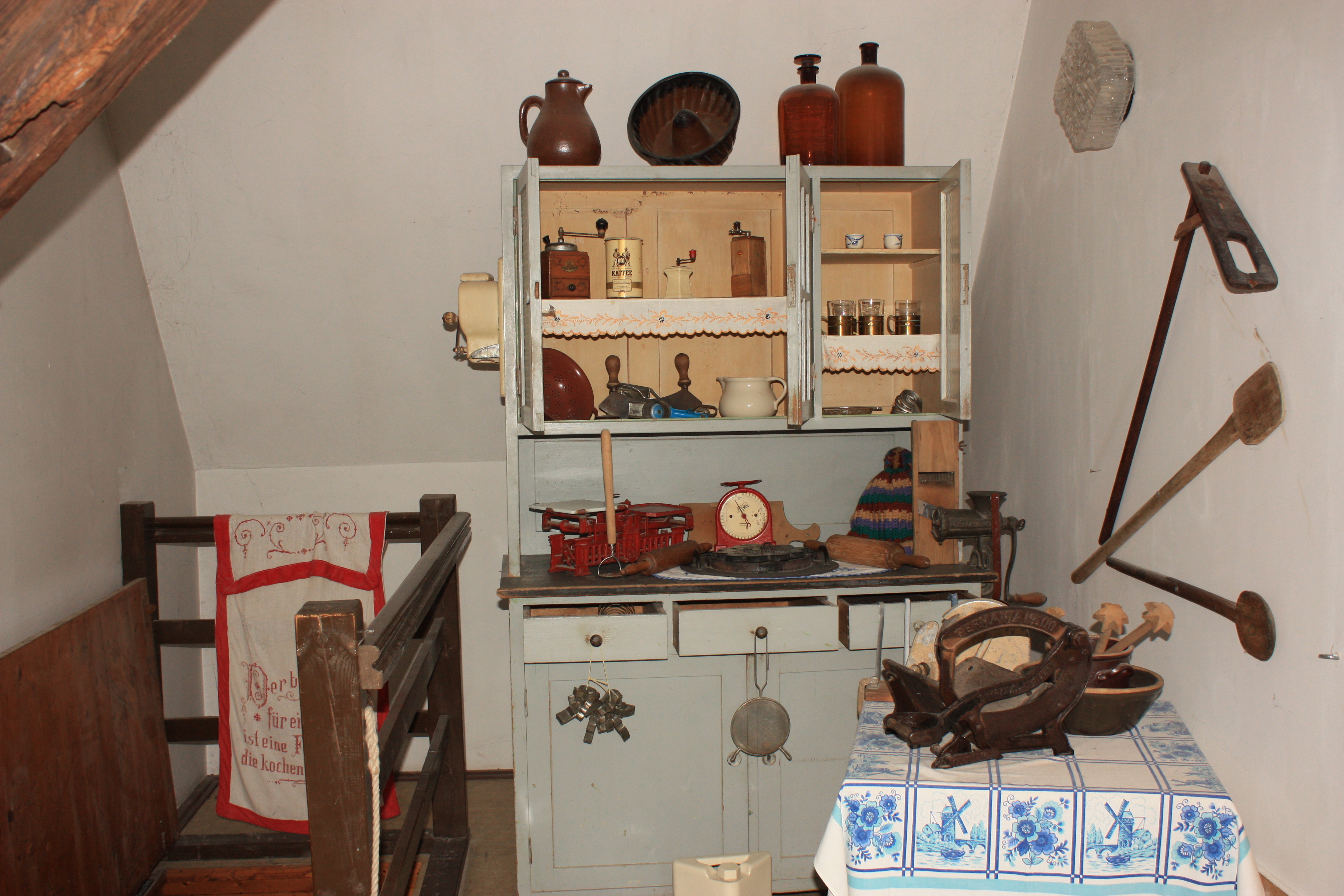 Ein kleines Zimmer mit einem Schrank voller Küchengeräte. Davor steht ein Tisch mit einer weiß-blauen Tischdecke und weiteren Gerätschaften. An der Wand hängen drei Holzgeräte.