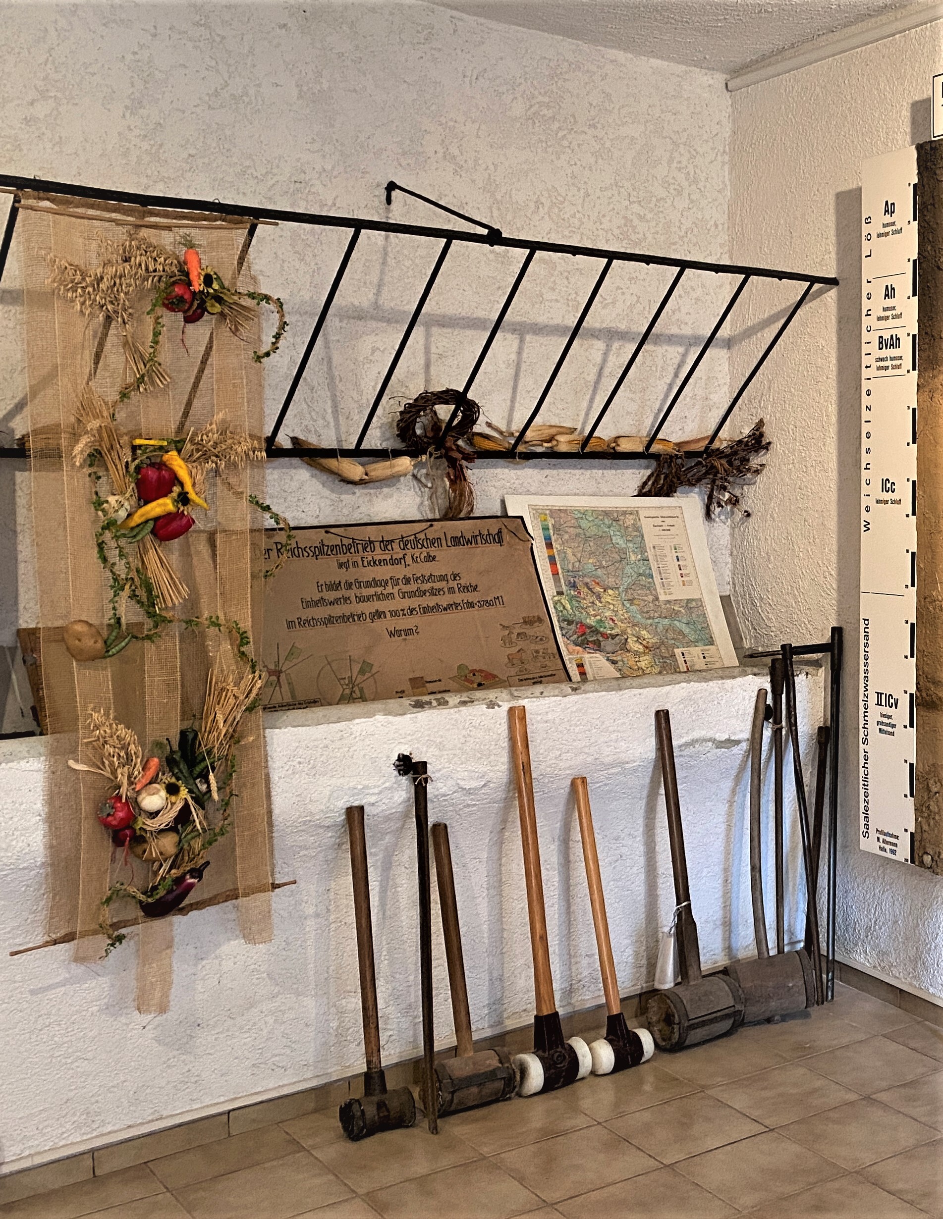 Die Werkzeuge der Bodenschätzer. An einer Wand stehen verschiedene Gerätschaften aus Holz an der Wand aufgereiht. Sie besitzen einen langen Stiel und einen Klotz am Ende. Darüber stehen eine Landkarte und ein Schild. Maiskolben hängen von einem Gitter herunter.
