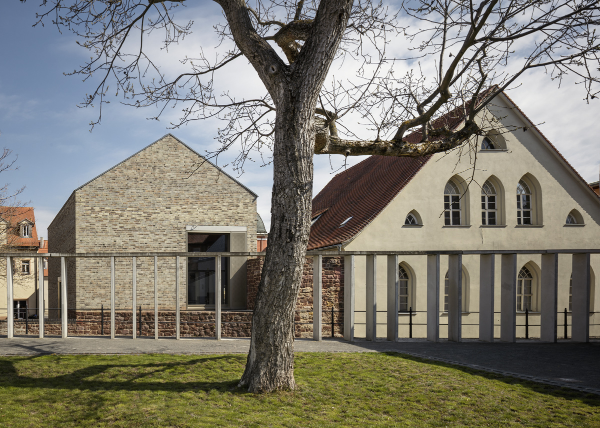 Luthers Geburtshaus. Im Vordergrund befindet sich eine grüne Wiese mit einem Baum ohne Blätter in der Mitte. Im Hintergrund stehen zwei graue Häuser aus Stein mit einem Dreiecksgiebel. Das linke Haus besitzt eine große Fensterfront.