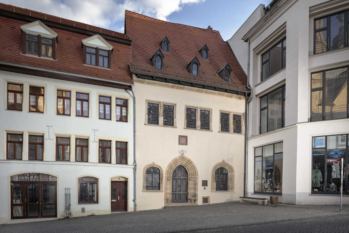 Luthers Sterbehaus. Das Museum wird von zwei Häuserfronten flankiert. Das linke Haus ist hellgrün und hat viele Fenster. Das rechte Haus ist weiß, größer und besitzt sehr große Fenster. Luthers Sterbehaus in der Mitte ist hellgelb gestrichen, der Eingang ist ein rundbogiges Portal und es besitzt nur eine Fensterreihe.