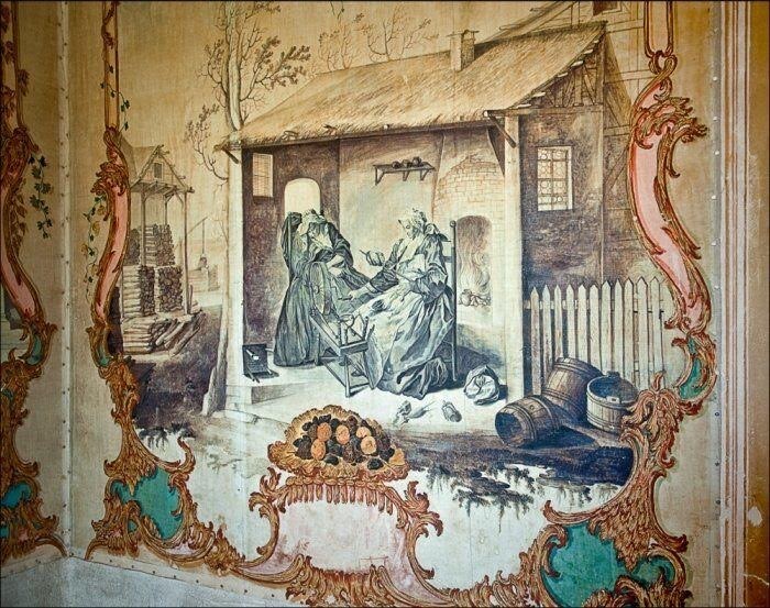 Restaurierte Gouache-Tapeten im Schloss. Das Motiv zeigt zwei Frauen vor einem Spinnrad. Die ältere Frau ist eingeschlafen und die jüngere arbeitet mit der Wolle in der Hand. Außerhalb des Hauses liegen Holzfässer und Holzstapel. Das Motiv wird von einer goldenen geschwungenen Rahmung eingefasst, die mit blauer und hellrosa Farbe ergänzt wurde.
