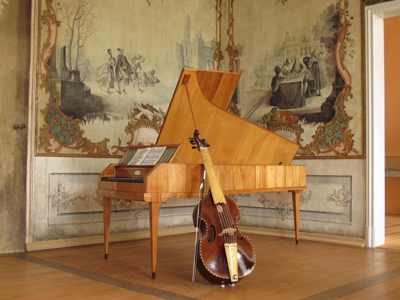 Gegen einen hellen Holzflügel ist ein großes Streichinstrument gelehnt. Im Hintergrund befinden sich die restaurierten Guache-Tapeten mit barocken Motiven.