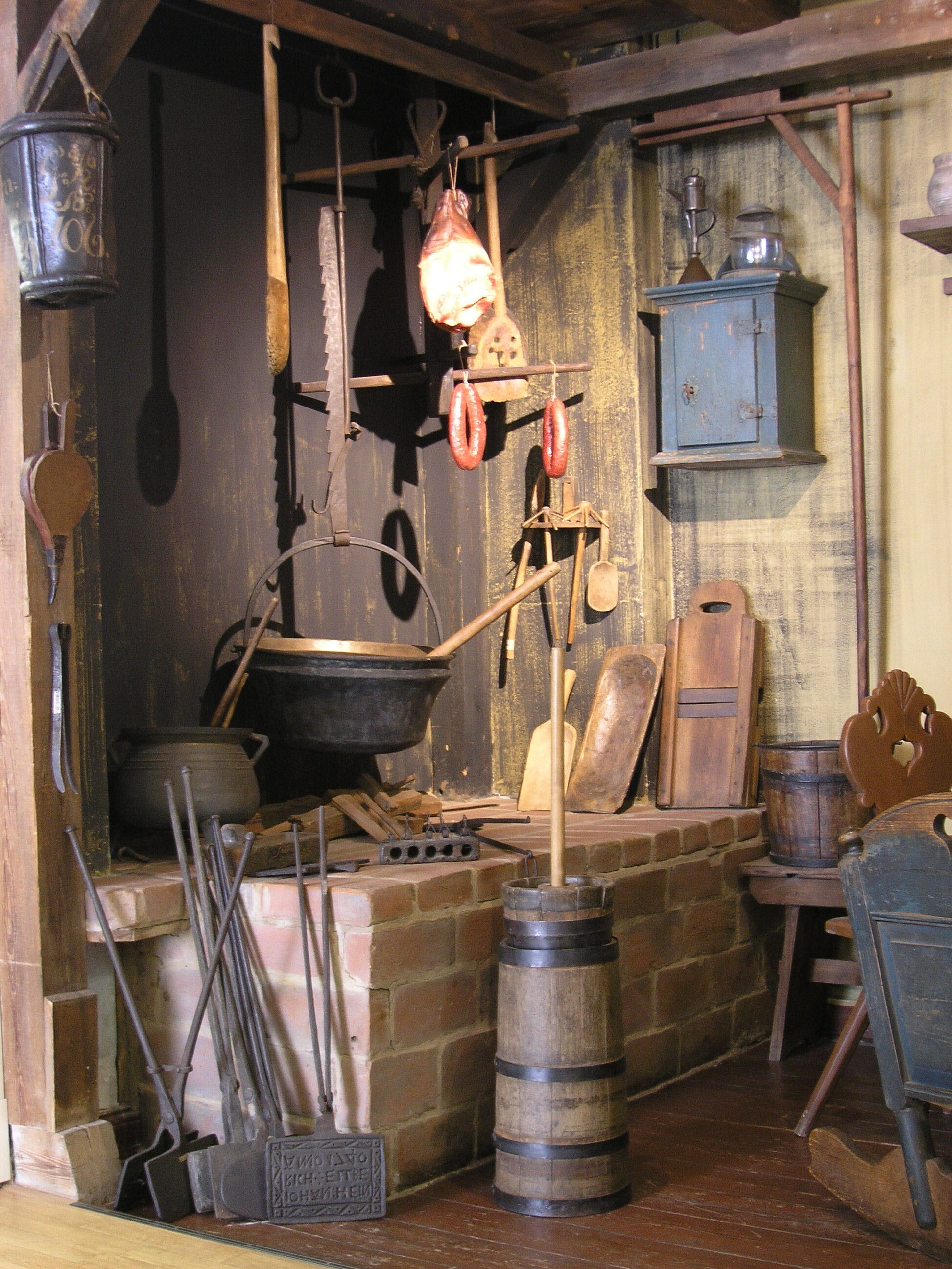 Schwarze Küche. Über einer Feuerstelle aus Backstein hängt ein schwarzer Kessel mit einer Kelle. Darüber wird Fleisch getrocknet. Einige Küchengeräte umgeben das Kochfeld.