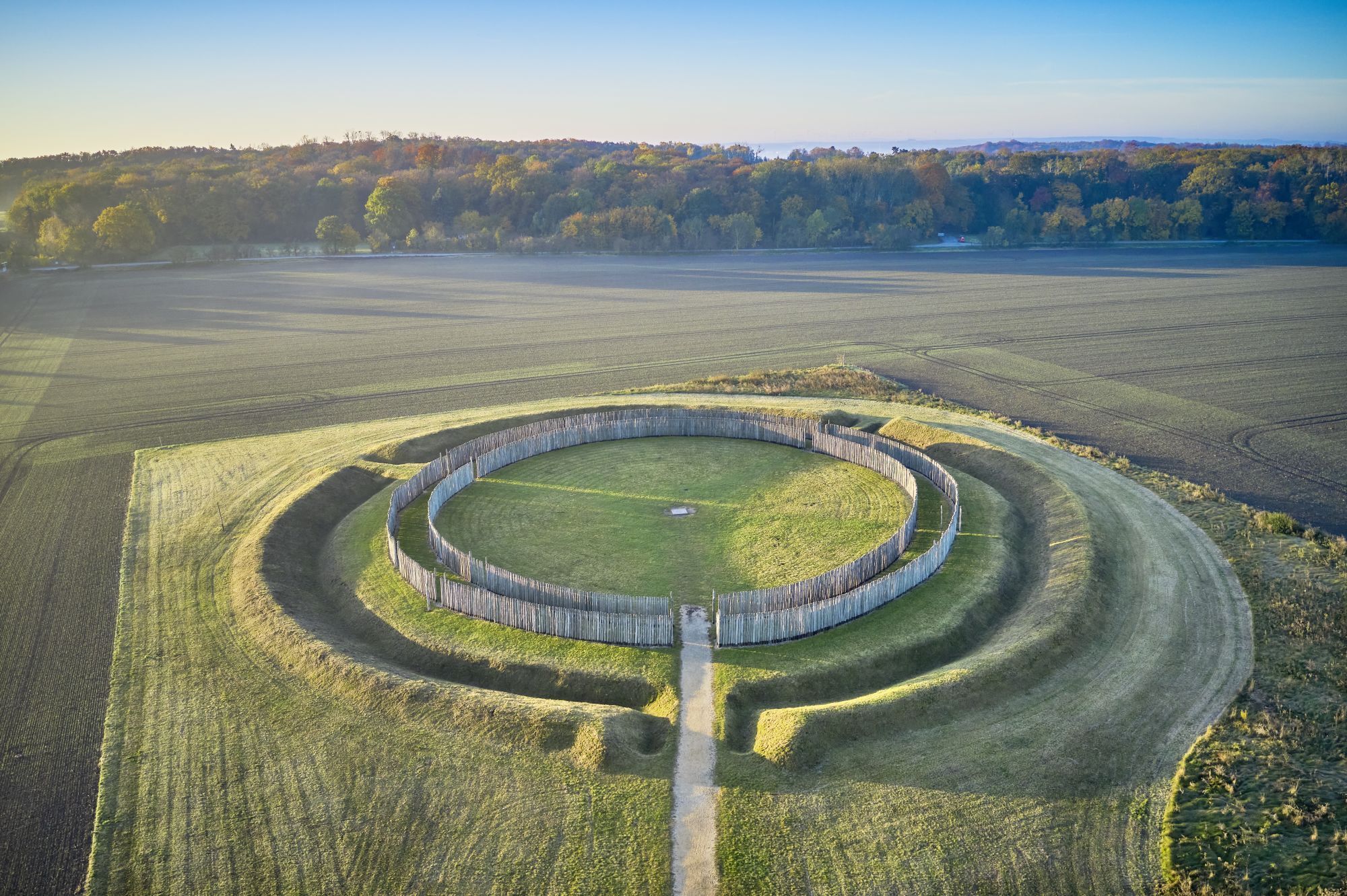 Die Rekonstruktion des Sonnenobservatoriums. Auf einem Feld ist eine große Fläche von Gras bedeckt. Ein schmaler Weg führt in das Innere eines Kreises, der aus Holzpfählen gebaut wurde. Um den Kreis wurde ein Graben gezogen.