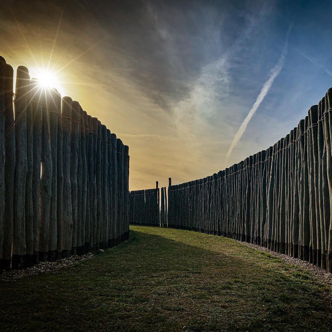 Sonnenobservatorium. Ein Grasweg führt um eine Kurve. Er wird von hohen Holzpfählen an beiden Seiten umgeben. Im Hintergrund ist der Sonnenuntergang zu sehen.