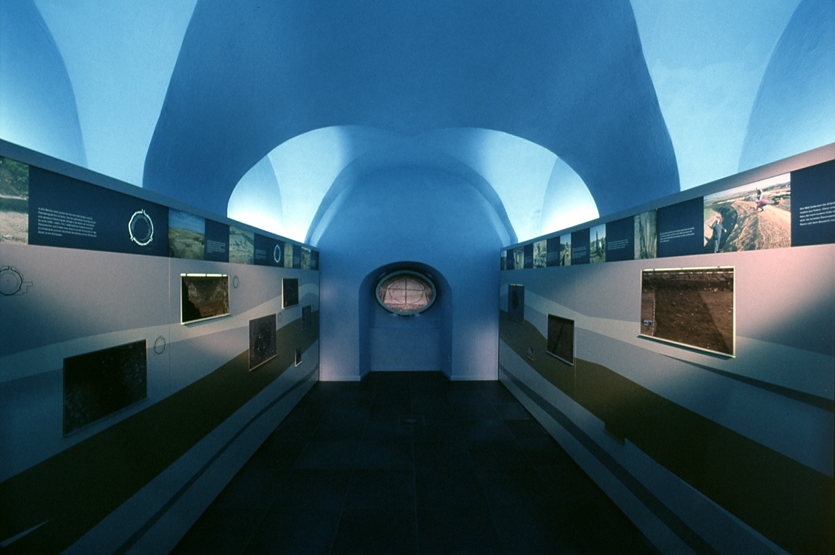 Blick in die Ausstellung des Informationszentrums. Ein blauer langgezogener Raum mit schmalen Informationstafeln an den Seiten.