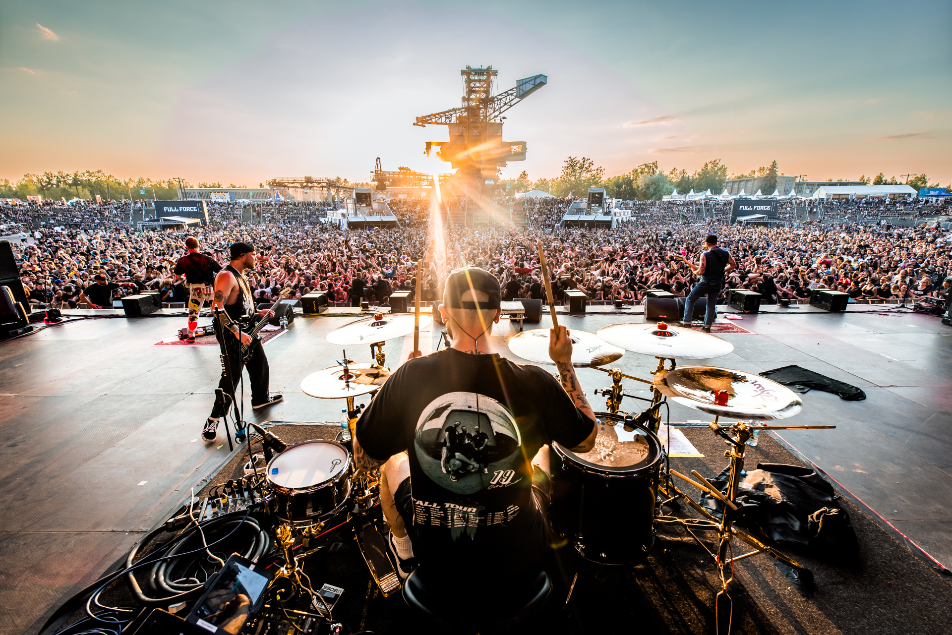 FULL FORCE 2023. Ein Schlagzeuger ist von hinten zu sehen. Hinter ihm stehen Bassisten, die sich zum vollen Publikum drehen. Im Hintergrund ist ein Braunkohle-Bagger zu sehen.