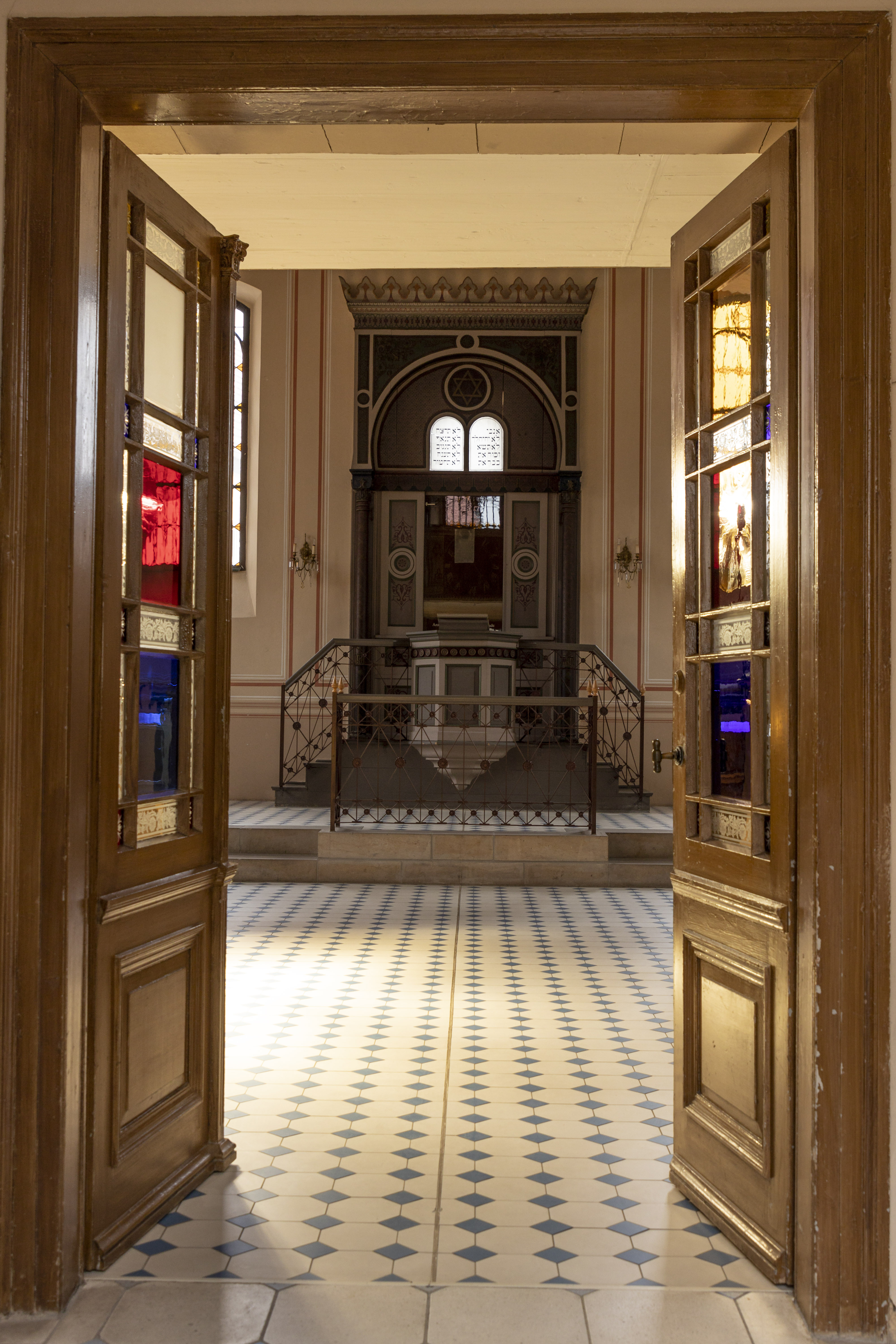 Zwei Türen mit Buntglas geben den Blick auf eine Bima frei. Der Bereich um die Bima ist grau und weiß gehalten. Zwei kleine Treppen an der Seite führen zum Podium. Der Boden ist weiß und blau gefliest.
