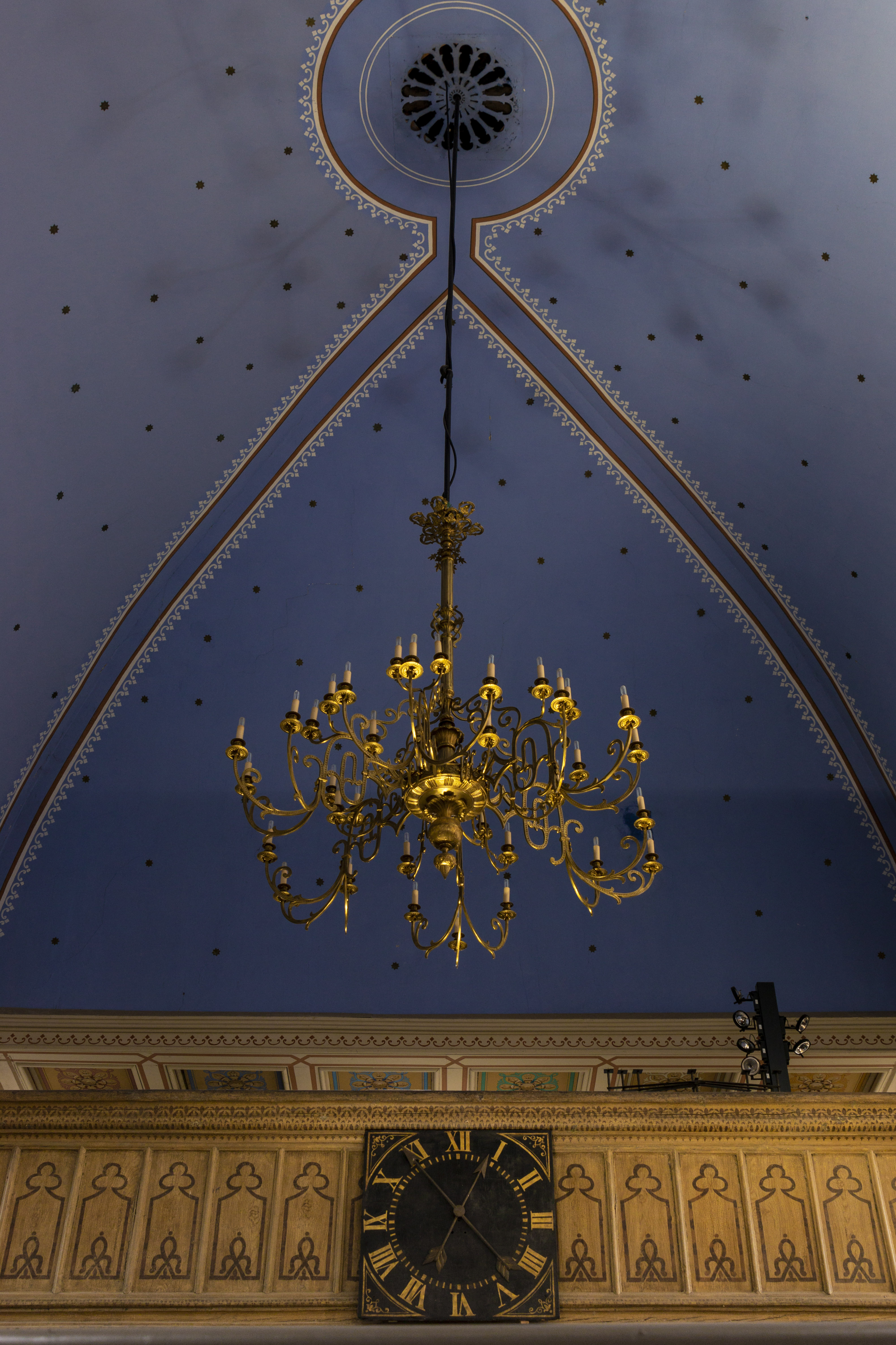 Ein goldener Kronleuchter hängt von einer blauen Decke, die mit schwarzen Punkten verziert ist. Auf einer Holzgalerie ist eine schwarze Uhr mit goldenen Ziffern angebracht.