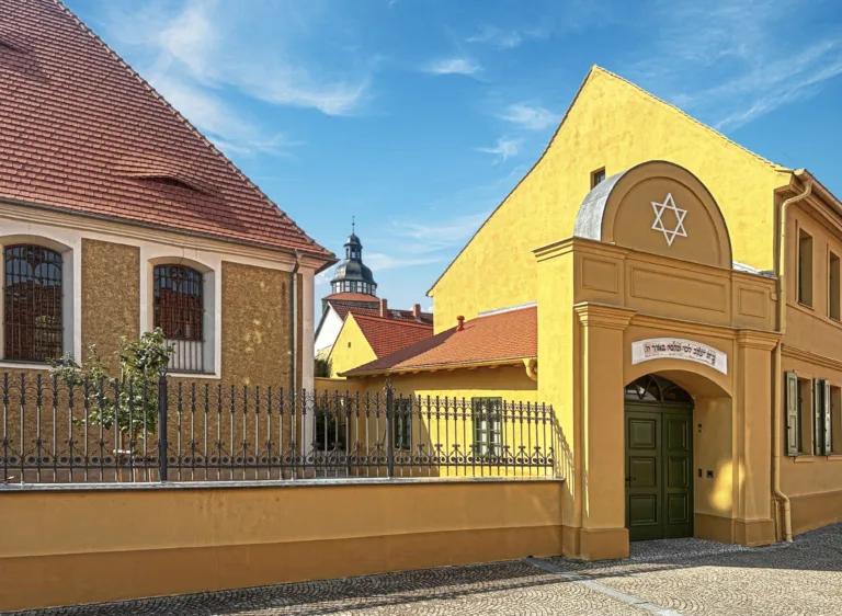 Eingangsbereich der Synagoge Gröbzig. Zwei gelbe Häuserfassaden und ein Kirchturm im Hintergrund sind erkennbar. Vor dem linken Haus befindet sich ein Zaun. Die linke Fassade gehört zu der Synagoge. Über dem Eingangsbereich befindet sich ein weißer Davidstern.
