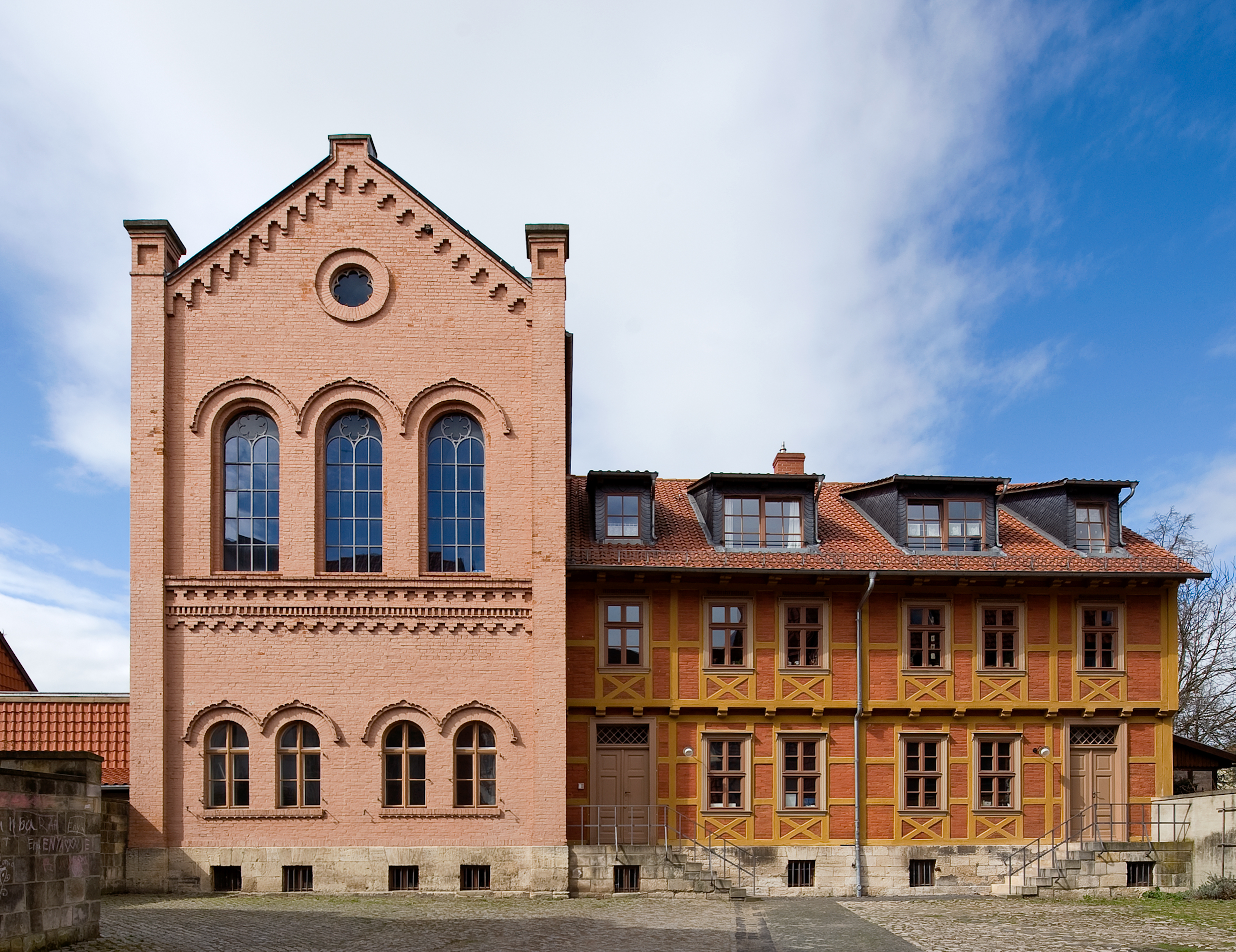Klaus bei Tag. Die Klaus ist ein jüdisches Lehrgebäude, das hier von Außen dargestellt ist. Es besteht aus einem höheren Bau mit einem Dreiecksgiebel und drei schmalen hohen rundbogigen Fenstern. Ein länglicher Anbau aus rotem und gelbem Fachwerk schließt sich an.